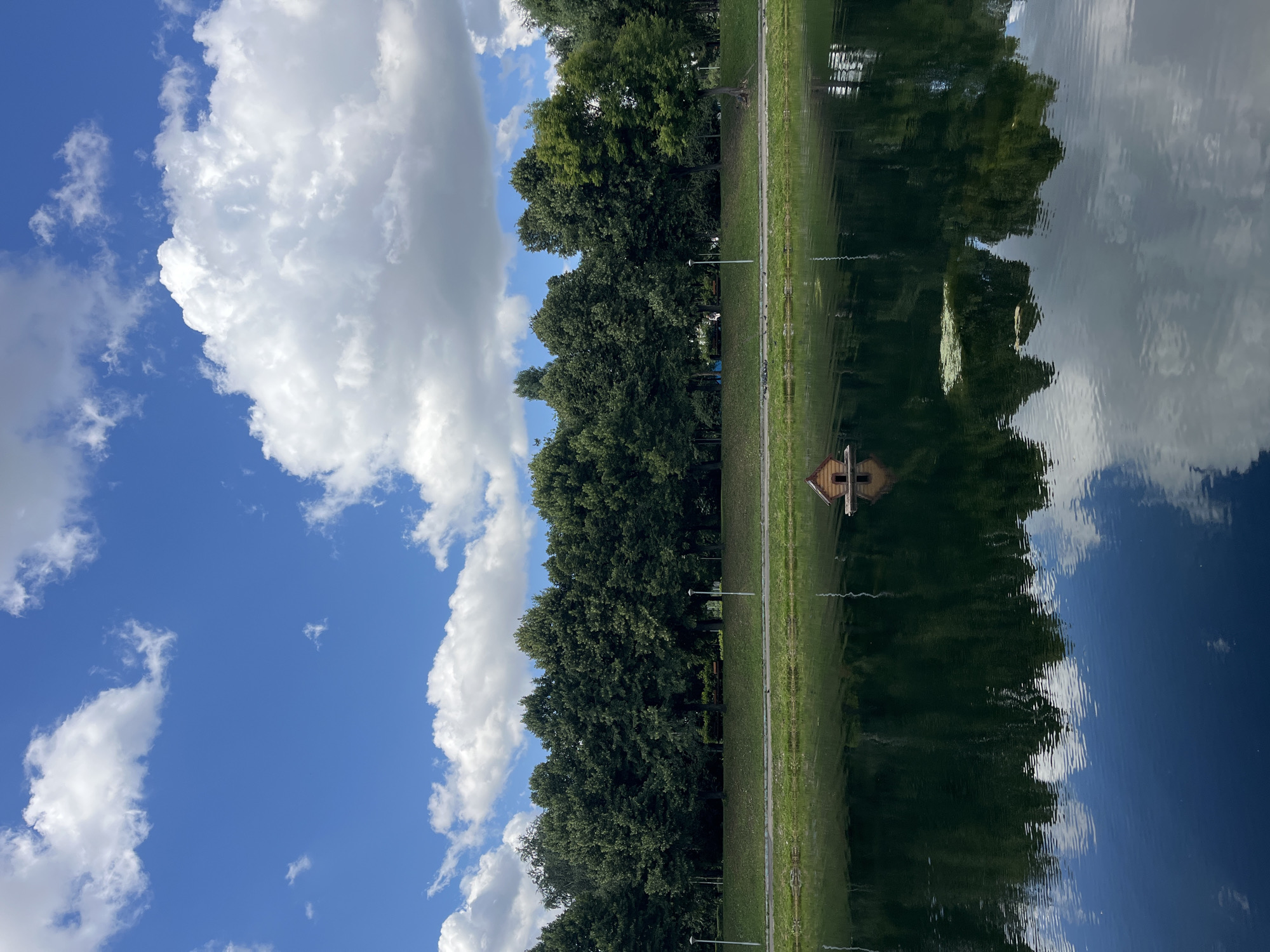 This is a vertically oriented, wide-angle shot of a park with a body of water reflecting the sky and trees. The scene is dominated by the reflection, with the sky occupying the upper half of the image and a line of trees bordering a grassy area to the left. The clouds in the sky are white and fluffy, contrasting with the deep blue sky.
In the reflection, the trees appear as dark silhouettes, while the sky's reflection shows streaks of white cloud and blue. A small, wooden structure, possibly a birdhouse or a miniature house, is visible on the edge of the water, also reflected. The grassy area to the left of the water is well-maintained, and a few streetlights are visible along the path bordering the trees. There are no people clearly visible in the image, although there might be some in the distance on benches in the park.
The overall impression is one of tranquility and nature. The time of day appears to be daytime, given the bright sunlight illuminating the clouds and the clear sky. The weather seems pleasant, with the calm water indicating a lack of strong wind. The setting is a park or a natural area with a lake or pond, likely in a suburban or rural location, given the presence of trees and manicured grass. There is no visible text in the image.