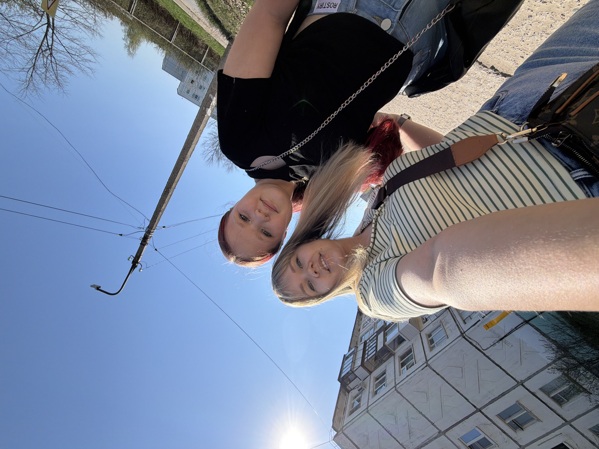 Two young women are posing for a photo outdoors. The photo is taken from a low angle, looking up at the women against a clear blue sky. The woman on the left has red hair and wears a black top, while the woman on the right has blonde hair and wears a striped shirt. A building with a grid pattern on its exterior is visible in the background. A street light and power lines are also in view. The location is likely Chaikovsky, Russia.