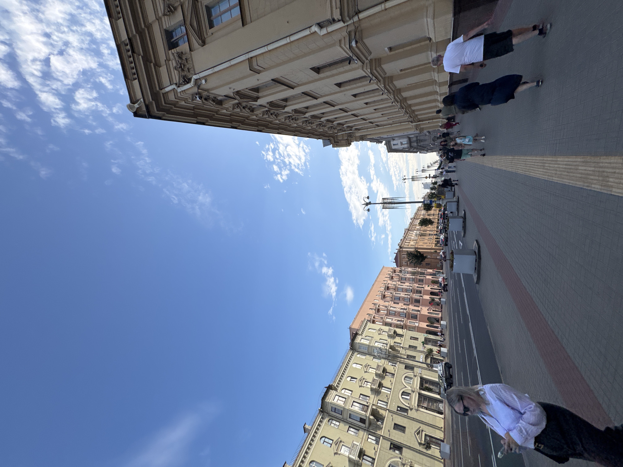 The image captures a street scene in Minsk, Belarus, featuring a wide sidewalk and a road with several vehicles. On the left, a row of colorful buildings with balconies lines the street. On the right, a large, ornate yellow building with numerous windows dominates the view. Several people are walking on the sidewalk, some are looking at their phones, and others are walking in groups. The sky is blue with scattered clouds. The perspective of the image is angled upwards, giving a sense of the grand scale of the architecture.