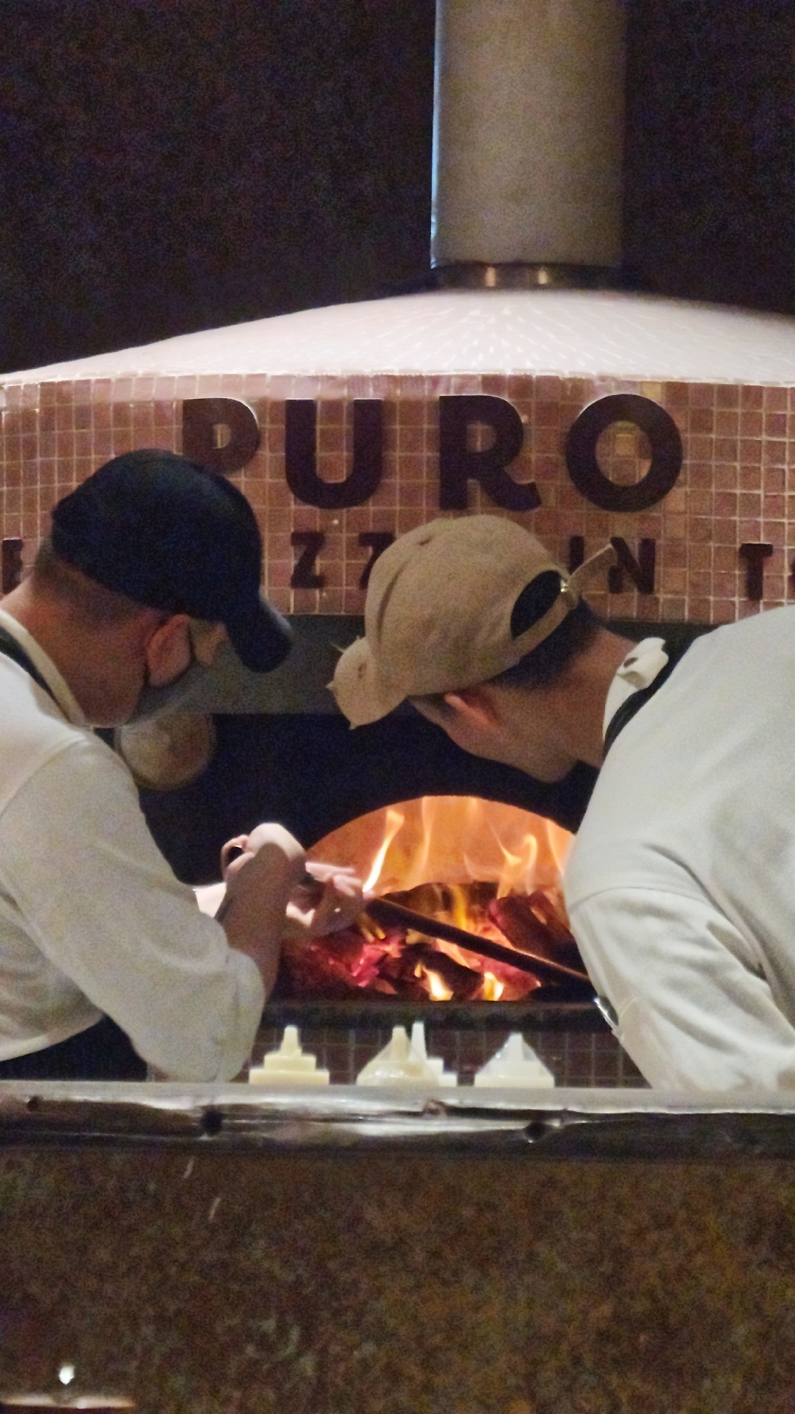 Two cooks are working in front of a wood-fired oven. The oven is marked "PURO" and has a tile exterior. One cook wears a black baseball cap and a face mask, and is using tongs to tend to the fire. The other cook wears a tan baseball cap. Both cooks are wearing white shirts. The image appears to be taken inside a restaurant in Hà Nội, Vietnam. In front of the oven are four small containers.