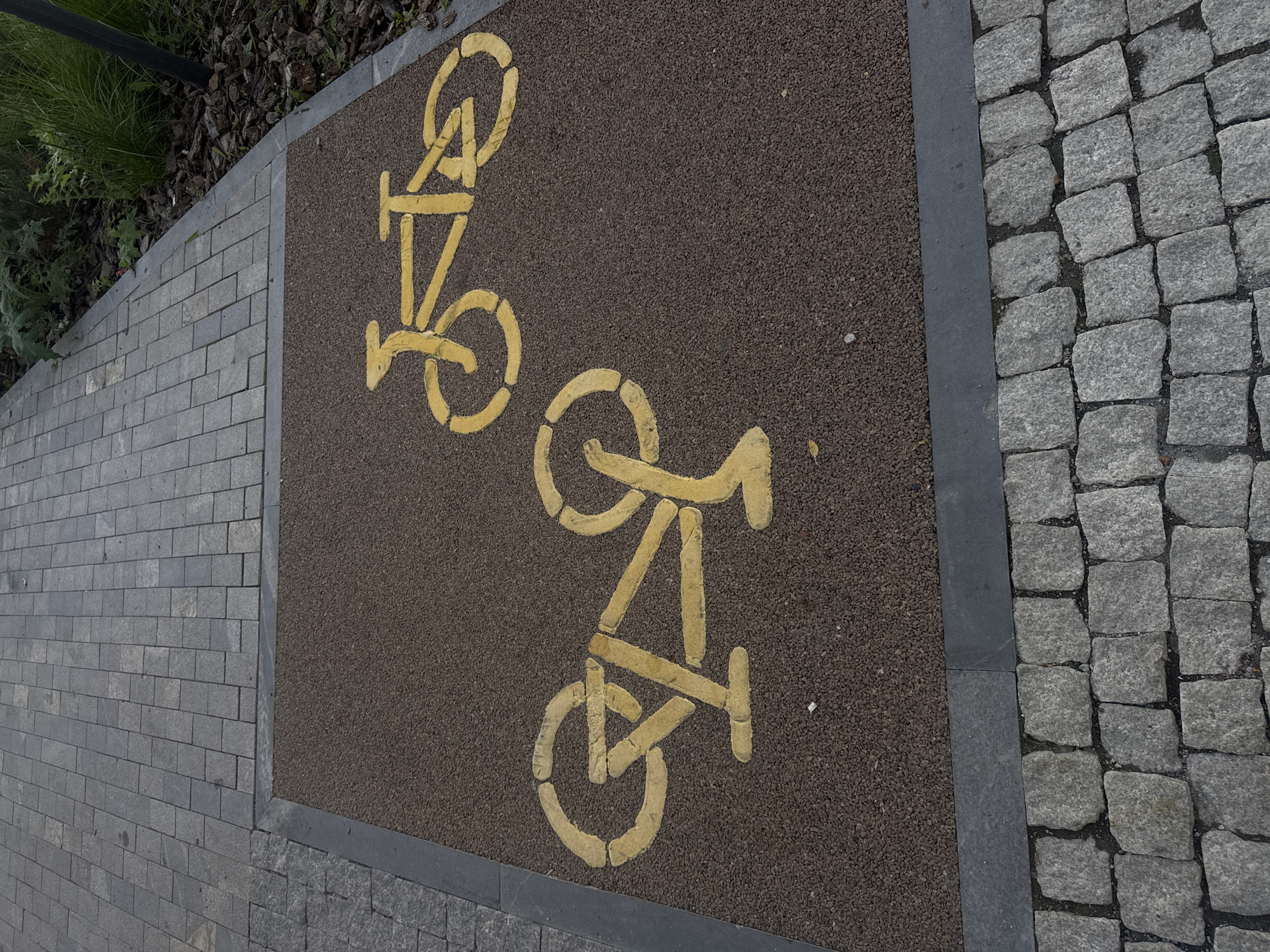 The image shows a close-up, overhead view of a bike path in Yekaterinburg, Russia. The path itself is made of a dark, granular material and is marked with two yellow stenciled bicycle symbols. One symbol is positioned higher and to the left, while the other is lower and to the right. The path is bordered on the left by grey rectangular paving stones and on the right by irregular grey cobblestones. In the upper left corner, a patch of dark mulch with some green grass and foliage is visible, suggesting the bike path is located in an outdoor urban environment, possibly a park or a dedicated street lane. The lighting suggests it is daytime, and the weather appears to be overcast, as there are no harsh shadows. There is no text visible in the image. The focus is on the graphic markings of the bike path and its paving.