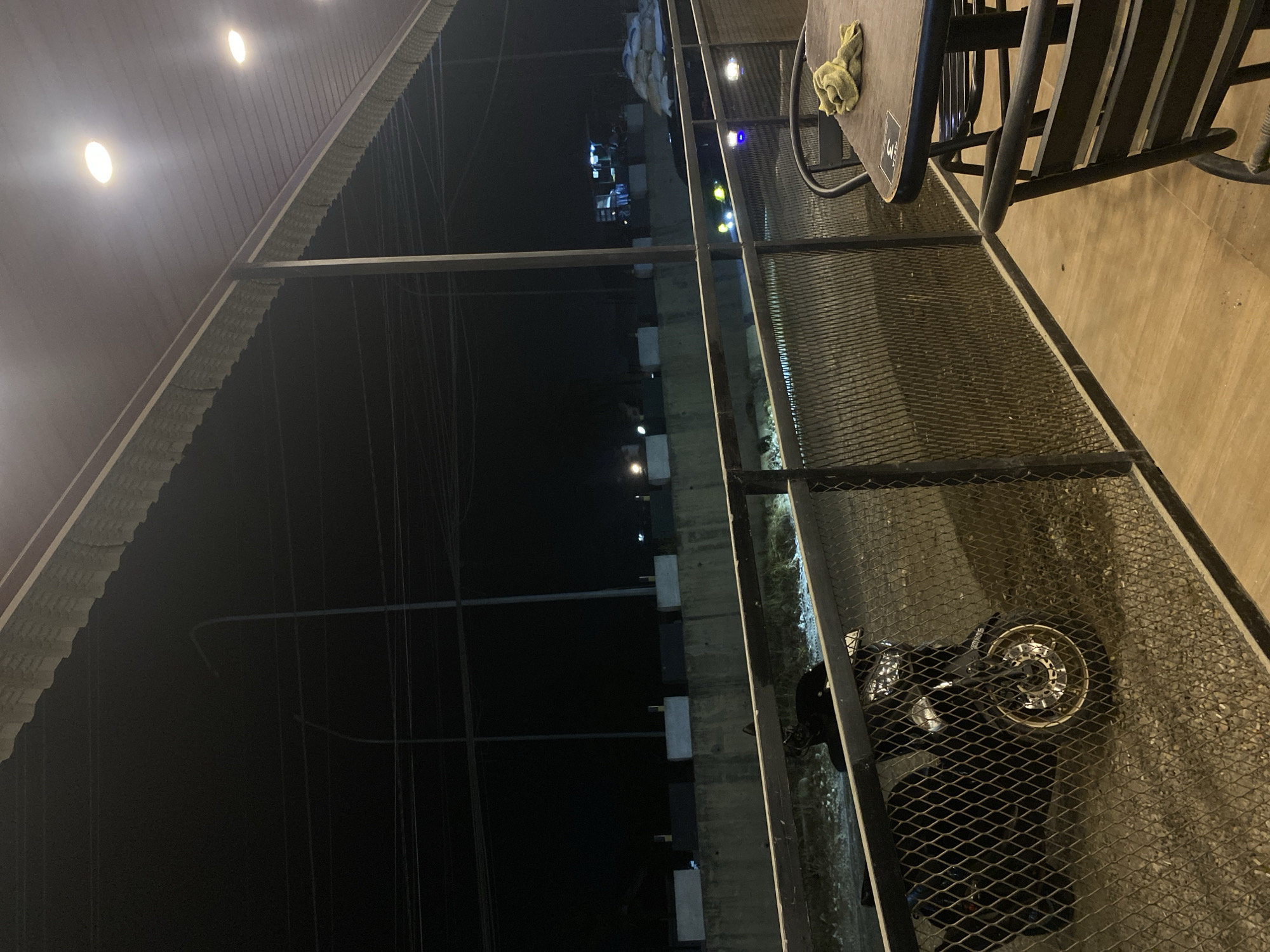 View from a covered patio area in Tanjongkapai, Indonesia, at night. A motorcycle is parked near a fence with a table and chairs on the patio. Streetlights are visible in the background.