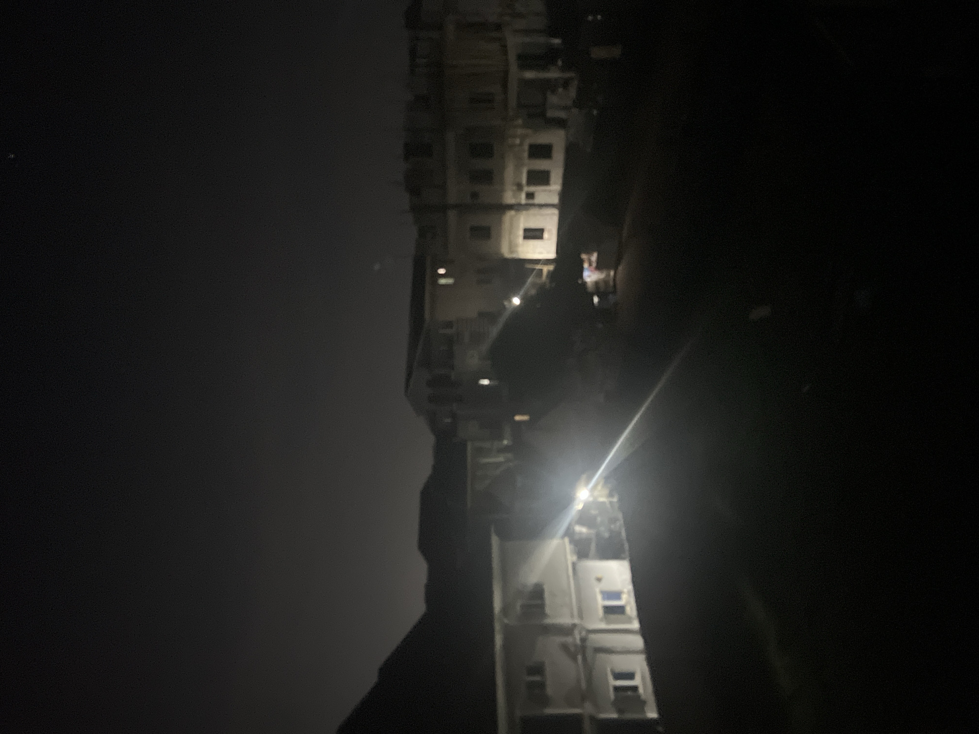 The image depicts a dark, night scene in Abuja, Nigeria, likely residential. A tall building is visible with multiple lit windows, suggesting activity inside. A bright light source is present, possibly a streetlight or from a window, casting a glare. The background is mostly dark, indicating low ambient light. There is a figure standing on what looks to be a balcony. The exact location within Abuja cannot be definitively determined from the image.