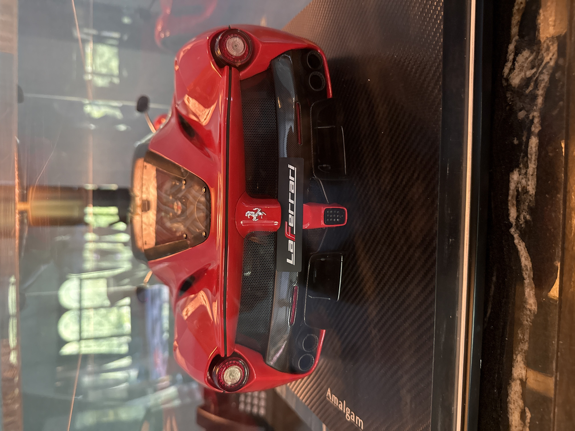 The image shows a red LaFerrari model car displayed within a glass case. The car is positioned on a carbon fiber patterned base. The name "LaFerrari" is visible on the rear of the car. The word "Amalgam" is printed near the bottom right corner of the base. Reflections of a building or window can be seen in the glass of the display case, suggesting the image was taken indoors. The environment appears to be a showroom or a collector's display. There is no direct evidence to pinpoint the exact location as Vila Verde, Portugal.