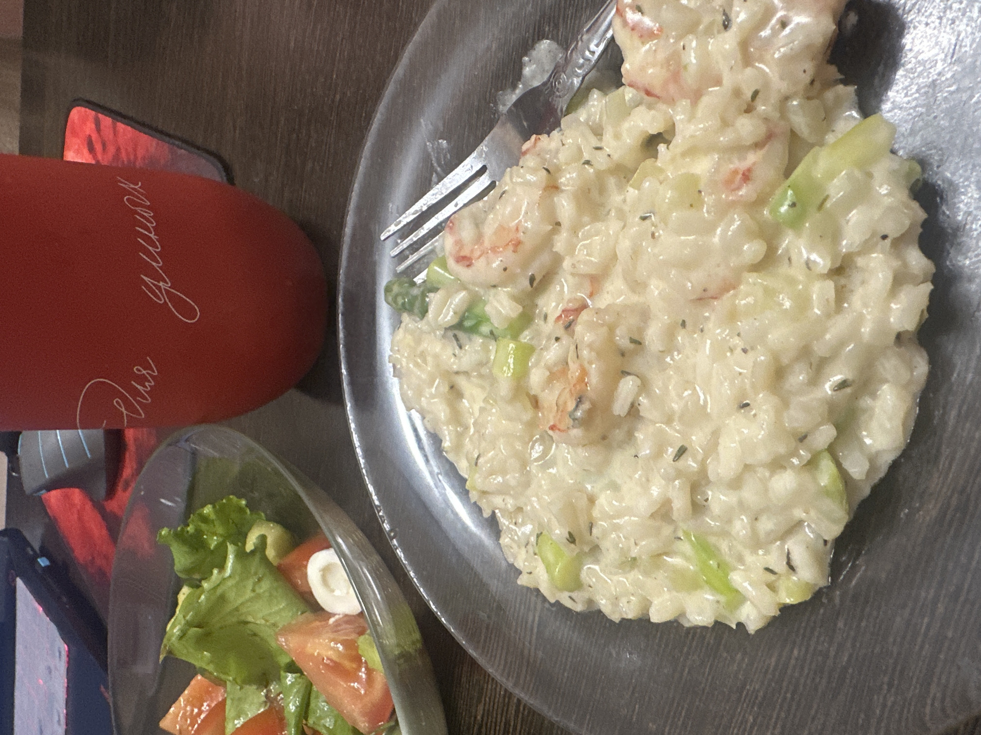 The image shows a close-up of a plate of shrimp risotto with asparagus, served on a dark, textured plate. A fork rests on the plate, with some risotto clinging to it. To the left of the risotto is a bowl of salad containing lettuce and tomatoes. In the background, a red tumbler with white cursive writing is visible, along with a computer mouse and a red mousepad, suggesting a desk setting. The overall impression is of a meal being consumed at a desk. There is no information in the image to determine its location in the city of Novoaltaysk, Russia.