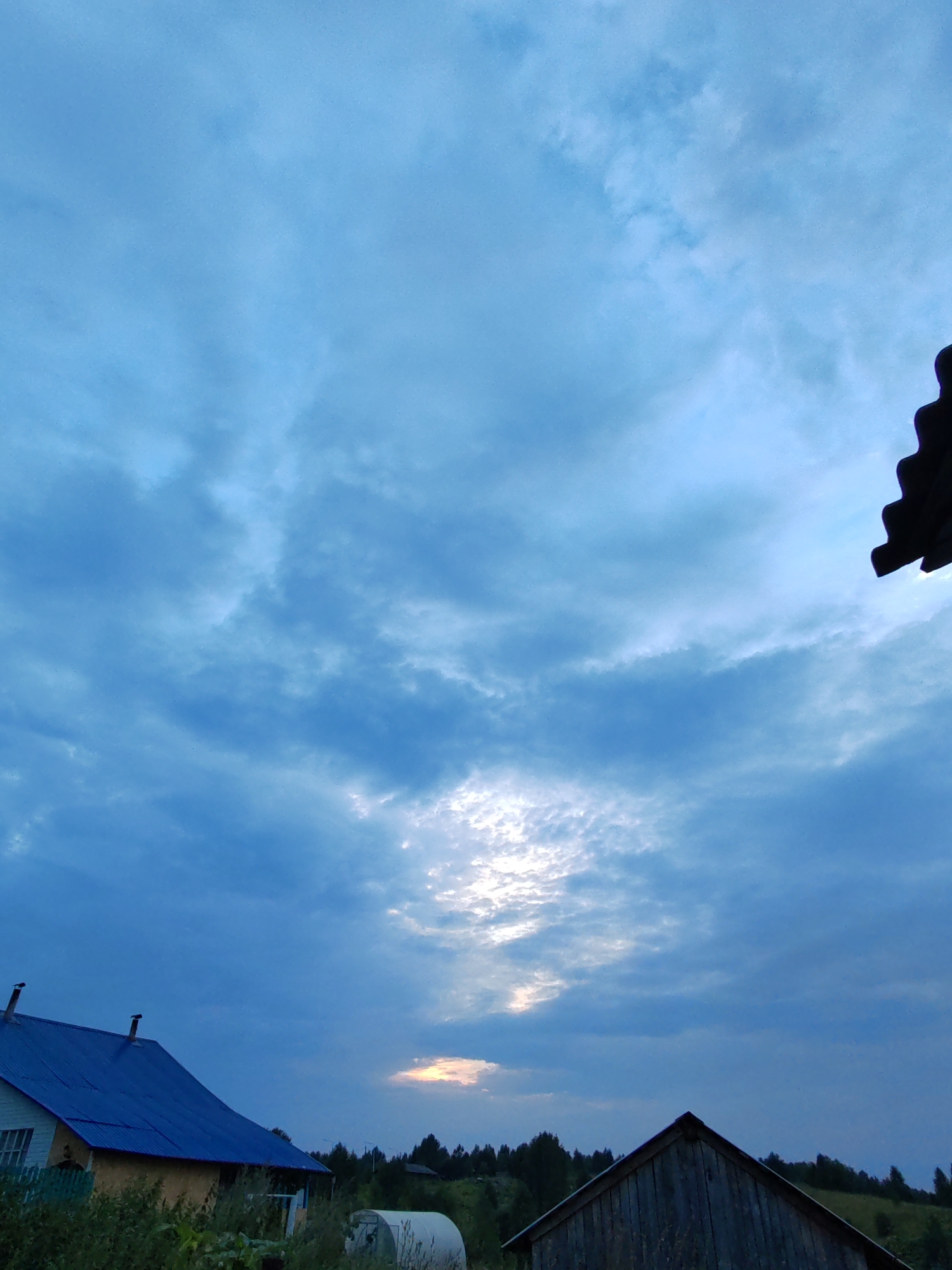 The image captures a twilight sky over a rural landscape, likely in the city of Chukhlem, Russia. The sky is filled with a dramatic array of clouds, predominantly in shades of blue and grey, with a bright patch of sunlight breaking through towards the center. In the foreground, two structures are visible. On the left is a building with a bright blue, corrugated metal roof and yellow siding. Its roof features two dark chimney-like protrusions. Green foliage encroaches on the lower part of this building. To the right, a darker, wooden structure with a peaked roof is silhouetted against the sky. Beyond these buildings, a dense line of trees forms a dark horizon, suggesting a forest or wooded area. The overall atmosphere is serene, indicative of the end of the day in a natural, undeveloped setting.