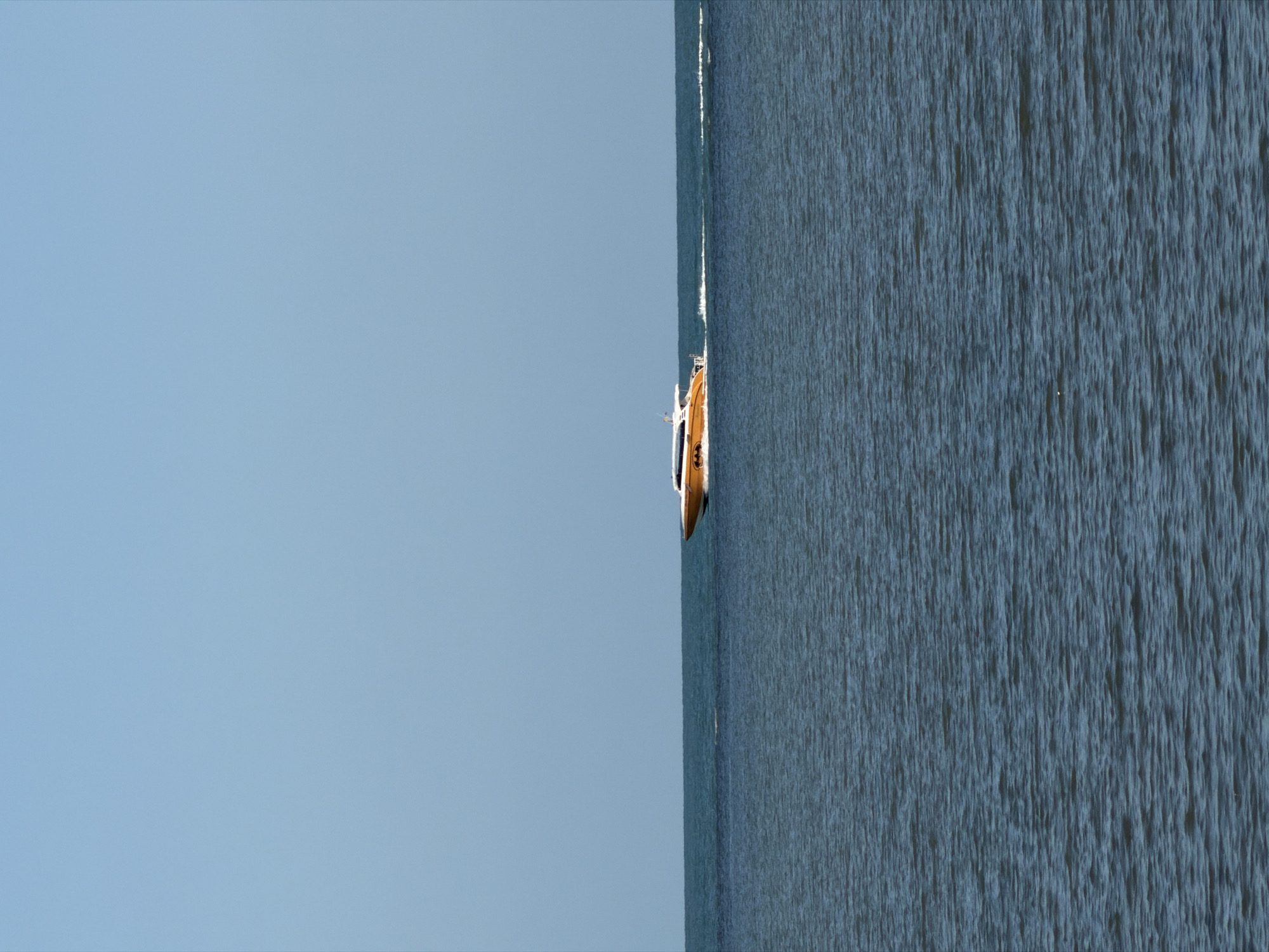 An orange and white speed boat is cruising on a calm, blue ocean under a clear, light blue sky. The boat is positioned on the right side of the image, moving from right to left, leaving a white wake behind it. The horizon line where the ocean meets the sky is clearly visible. The image appears to be taken at sea, likely in the waters surrounding Ko Samui District, Thailand, given the tropical, serene setting. There are no people visible on the boat.
