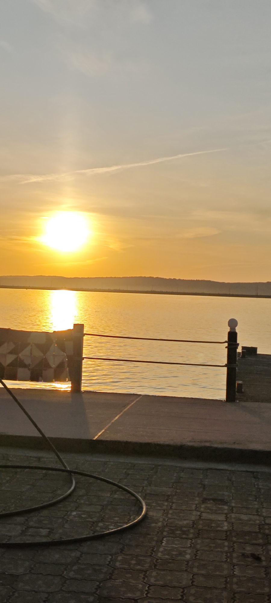 The image depicts a sunset scene from what appears to be a waterfront location. The sun is setting over a body of water, creating a bright, golden reflection on the water's surface. In the foreground, there is a section of a paved area, possibly a walkway or pier. Part of a barrier or railing is visible, constructed from posts connected by ropes. There is also a bench-like structure present. A black hose is partially visible in the foreground on the lower left. The location is identified as Chaikovsky, Russia, although there are no specific elements to confirm this.