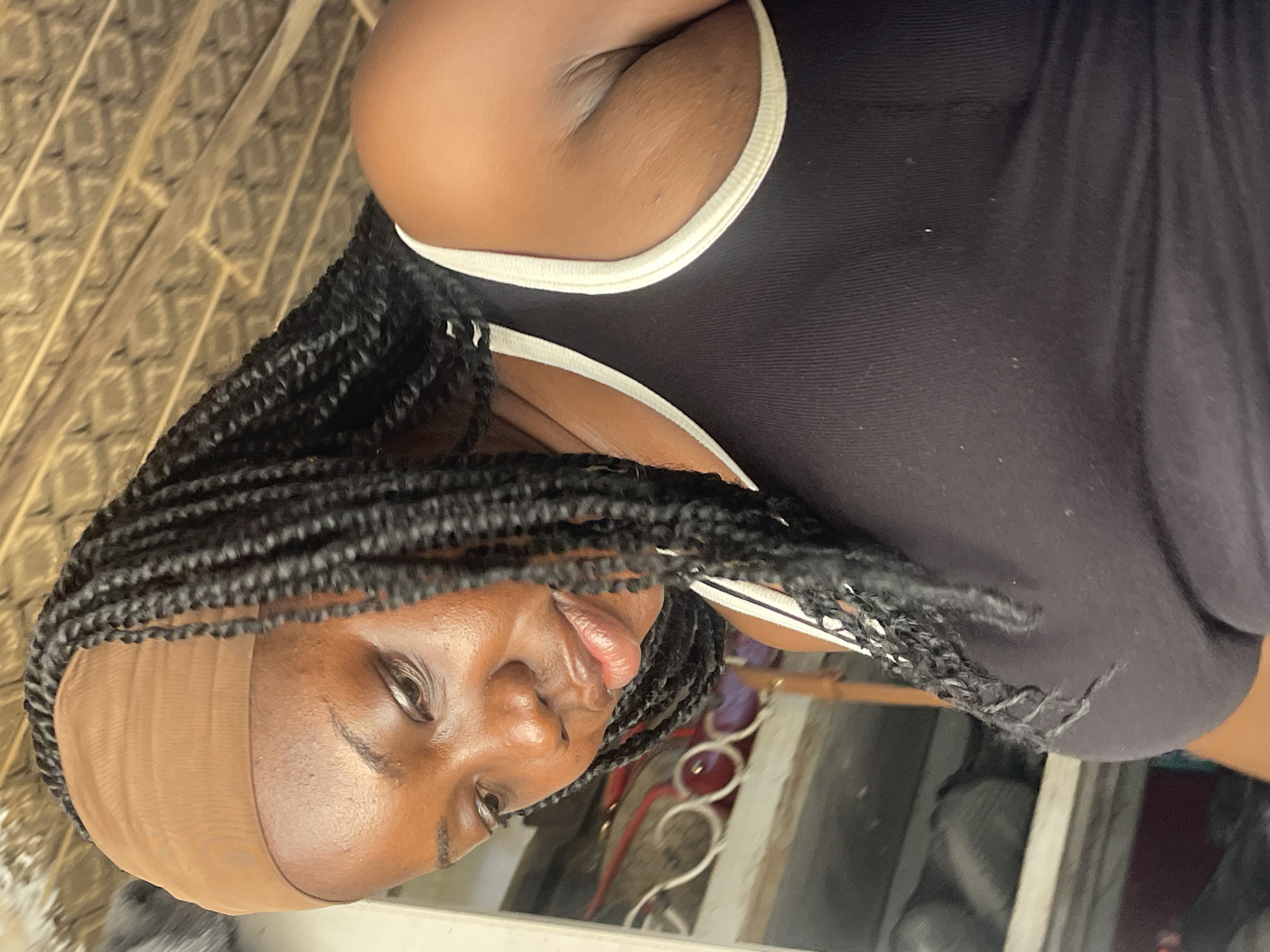 A medium shot captures a Black woman from the chest up, looking away from the camera. She has dark skin and is wearing a black tank top with white trim. Her hair is styled in long, dark braids that fall over her shoulders. A light brown wig cap is visible on her forehead. The background is somewhat blurred, but hints at an indoor setting with what appears to be a beige patterned wall or screen to her left and a glimpse of a reflective surface, possibly a mirror, below her. Behind her and to the right, there are hints of objects and possibly furniture in a dimly lit area, with some red accents. The lighting suggests it could be daytime, but the indoor setting makes it difficult to determine the exact time of day or weather conditions. The woman's expression is neutral, and the overall mood of the image is casual and relaxed. There is no visible text in the image.