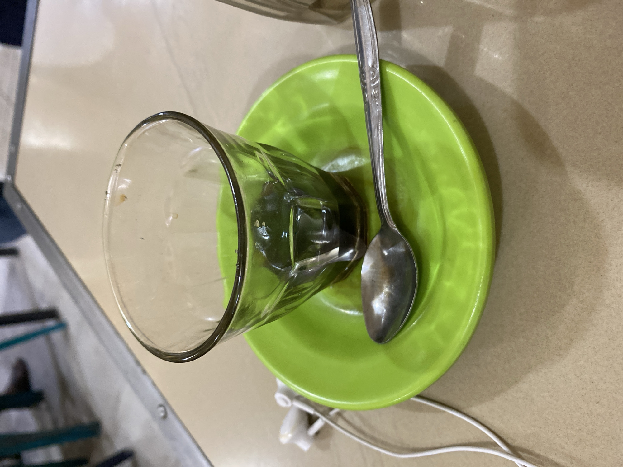 A close-up shot focuses on a partially filled glass of dark liquid, likely coffee, resting on a bright green saucer. A silver spoon is placed across the saucer. To the left of the saucer, a pair of white earphones with their white cord are visible. The scene appears to be set on a light brown tabletop. In the blurred background, glimpses of people's legs and the legs of chairs can be seen, suggesting an indoor setting like a cafe or restaurant. The overall impression is one of a paused moment, perhaps a break. Without additional information or context, it is not possible to definitively pinpoint the exact location in Lhokseumawe, Indonesia.