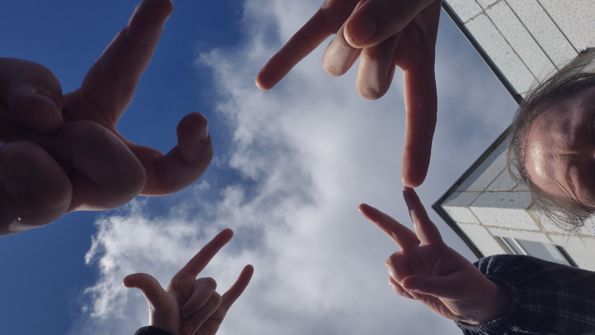 This low-angle photograph captures a playful interaction involving multiple hands and a partial view of a person, framed against a bright blue sky with scattered white clouds. Four hands are prominently featured in the foreground, positioned around the edges of the frame and pointing towards the center. Two hands, one in the top-left and another in the bottom-left, are making the "rock and roll" horn gesture (index and pinky fingers extended). The hand in the top-right and another in the bottom-right (near the person's head) are both forming a "peace" sign (index and middle fingers extended in a V-shape).
On the far right side of the image, the upper portion of a person's head is visible, including their forehead, part of an eye, and dark hair, suggesting they are looking down into the frame. The person appears to be wearing a dark plaid or checked shirt. In the background, a modern, light-colored building with a grid-like pattern on its facade can be seen, indicating an urban or built-up environment. The strong sunlight casts reflections on the person's forehead, suggesting the photograph was taken during bright daylight, likely midday or early afternoon, under fair weather conditions. The overall mood is casual and lighthearted. There is no visible text in the image. The location is Chaikovsky, Russia.