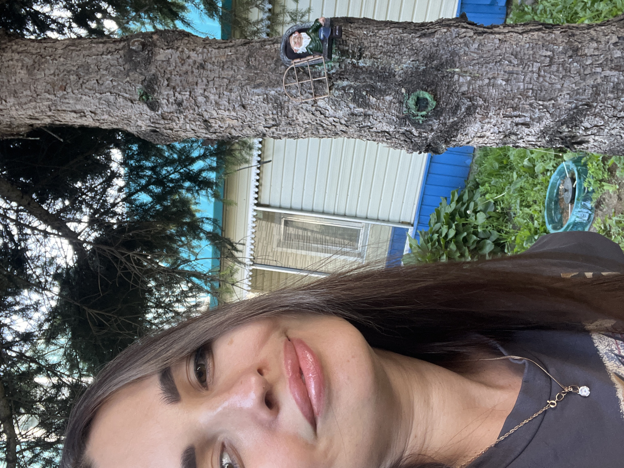 A woman is taking a selfie outdoors, with a tree and a building in the background. The woman, who appears to be in her late 20s or early 30s, has long brown hair and is wearing a dark t-shirt and a gold necklace with a diamond pendant. She is smiling and her lips are covered in lip gloss. The tree is a large, mature conifer with rough bark. A small gnome figurine is attached to the tree trunk. The building in the background is a single-story structure with light-colored siding and a blue base. The scene is set in a grassy, natural environment, possibly a yard or park.
