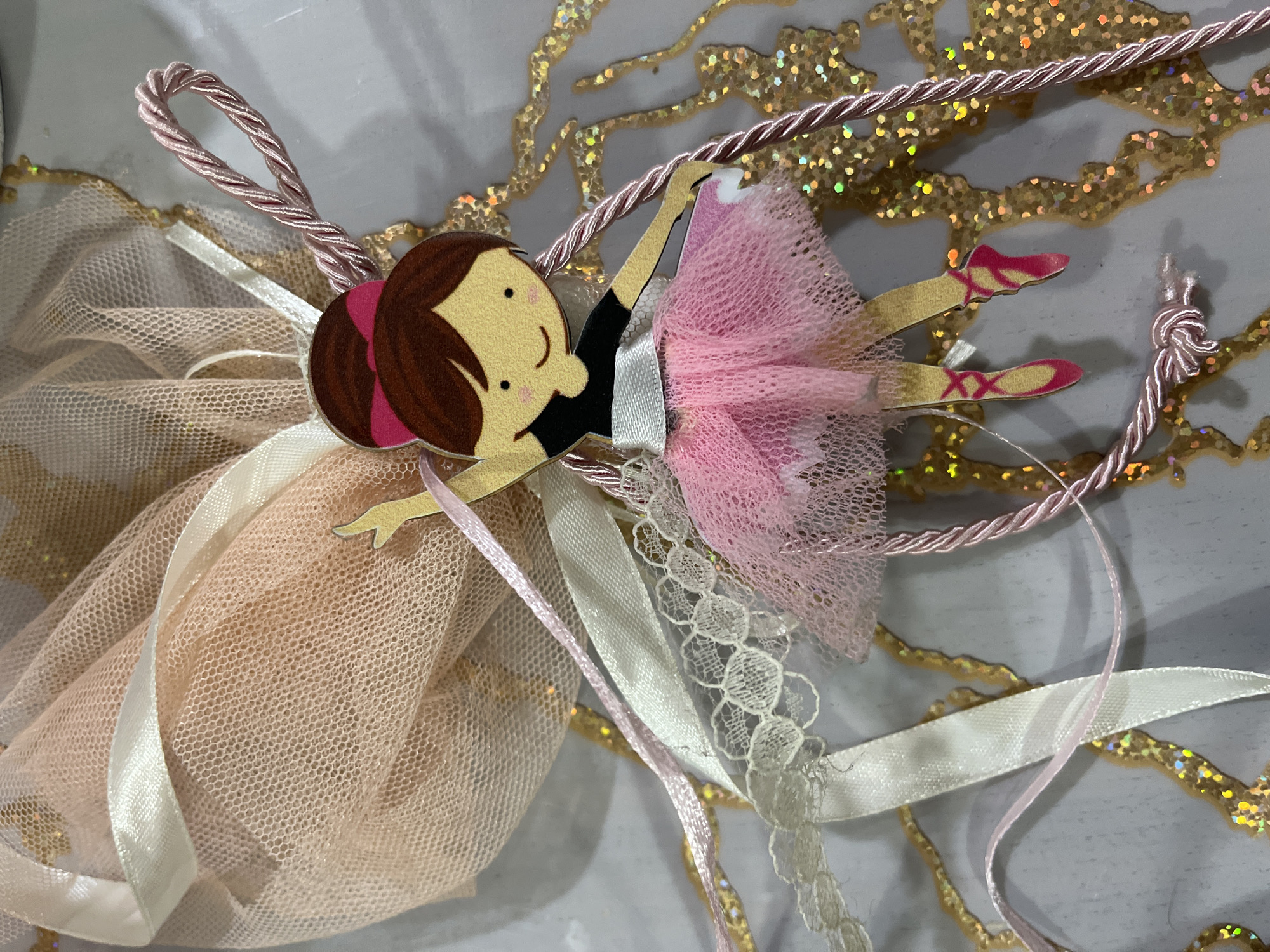 The image displays a decorative item featuring a ballerina motif, positioned on a light-colored surface with abstract gold glitter patterns. The central subject is a flat, cut-out figure of a ballerina with light skin, brown hair styled in a bun with a pink bow, a black short-sleeved top, and a white sash. A layered pink tulle skirt forms her tutu. She wears pink pointe shoes. The figure is depicted in a dynamic ballet pose with arms and legs extended, and a simple smiling face.
Attached above and to the left of the ballerina is a gathered bundle of light pink/peach sheer netting, secured by multiple ribbons. These include a wide white satin ribbon, a thinner light pink satin ribbon, and a white lace ribbon featuring a repeated circular pattern.
A twisted pink cord is integrated into the decoration. It forms a loop in the upper right portion of the image and extends downwards, appearing to be knotted or tied into a tassel near the bottom left, below the ballerina's feet.
The background surface is predominantly light gray or off-white, adorned with irregular, organic shapes composed of shimmering gold glitter. The glitter exhibits iridescent reflections. The lighting is bright and uniform. The location is unknown and not discernible from the image.