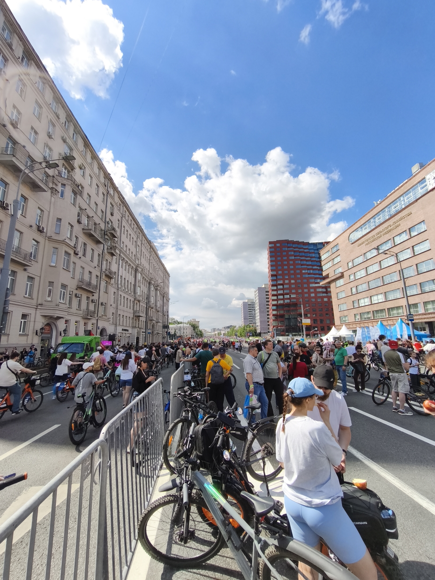 The image depicts a bustling street scene in Moscow, Russia, filled with people and bicycles. Buildings line the sides of the street. The building on the right side of the image is the Ministry of Agriculture of the Russian Federation, identifiable by the inscription on the building's facade. The sky is bright blue with scattered clouds. The street is crowded with people, many of whom are riding or standing with bicycles. A metal barrier runs along the edge of the street. People are interacting in small groups.