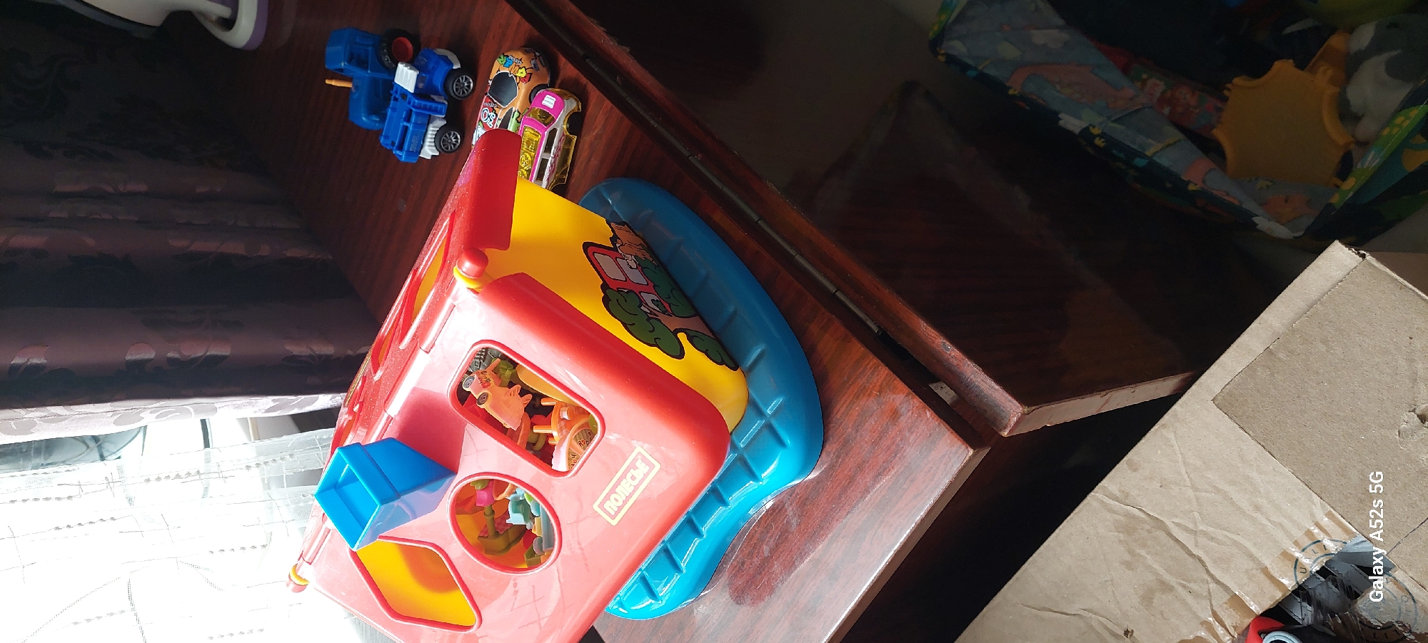The image shows a wooden table with several toys. On the left side of the table is a red and yellow toy house with various shapes to sort. Inside the toy house, there are small toy pieces. A blue plastic piece is on top of the toy house. To the right of the toy house are three toy vehicles: a blue tractor, a blue truck, and a colorful car. The table is in front of a window with sheer curtains and darker patterned curtains. The overall scene appears to be indoors, likely in a child's room or play area. There is also a cardboard box in the foreground.
