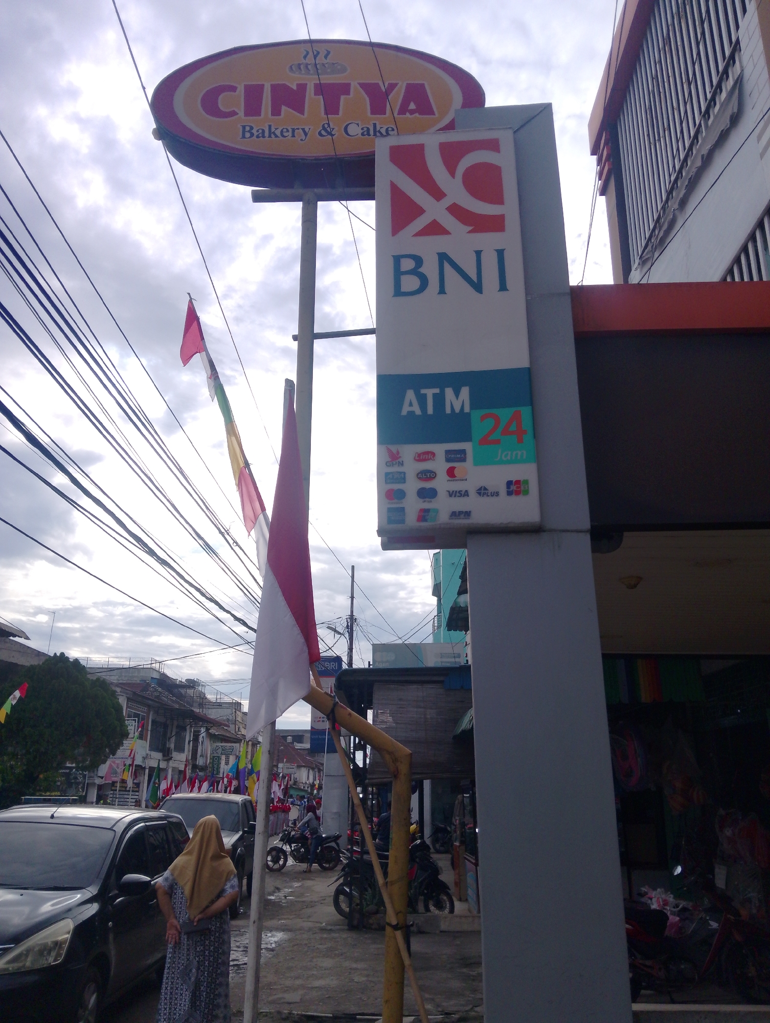 The image depicts an outdoor scene in Serbelawan, Indonesia. In the foreground, a woman wearing a tan hijab and a patterned dress stands with her back to the camera, facing a street. To her left is a black car, and behind her, another dark vehicle. The street is lined with buildings and appears to be a commercial area.
Dominating the upper portion of the image is a large, oval sign with "CINTYA Bakery & Cake" written on it. Adjacent to this, a vertical sign displays the "BNI" logo and information about an "ATM 24 Jam" (ATM 24 Hours), along with various credit card logos. Indonesian flags are visible, with a red and white flag prominently positioned near the ATM sign.
The sky is overcast with clouds. The environment suggests a typical day in a town or city, with street activity and signage indicating local businesses and services. The presence of multiple flags hints at a possible local celebration or national event.