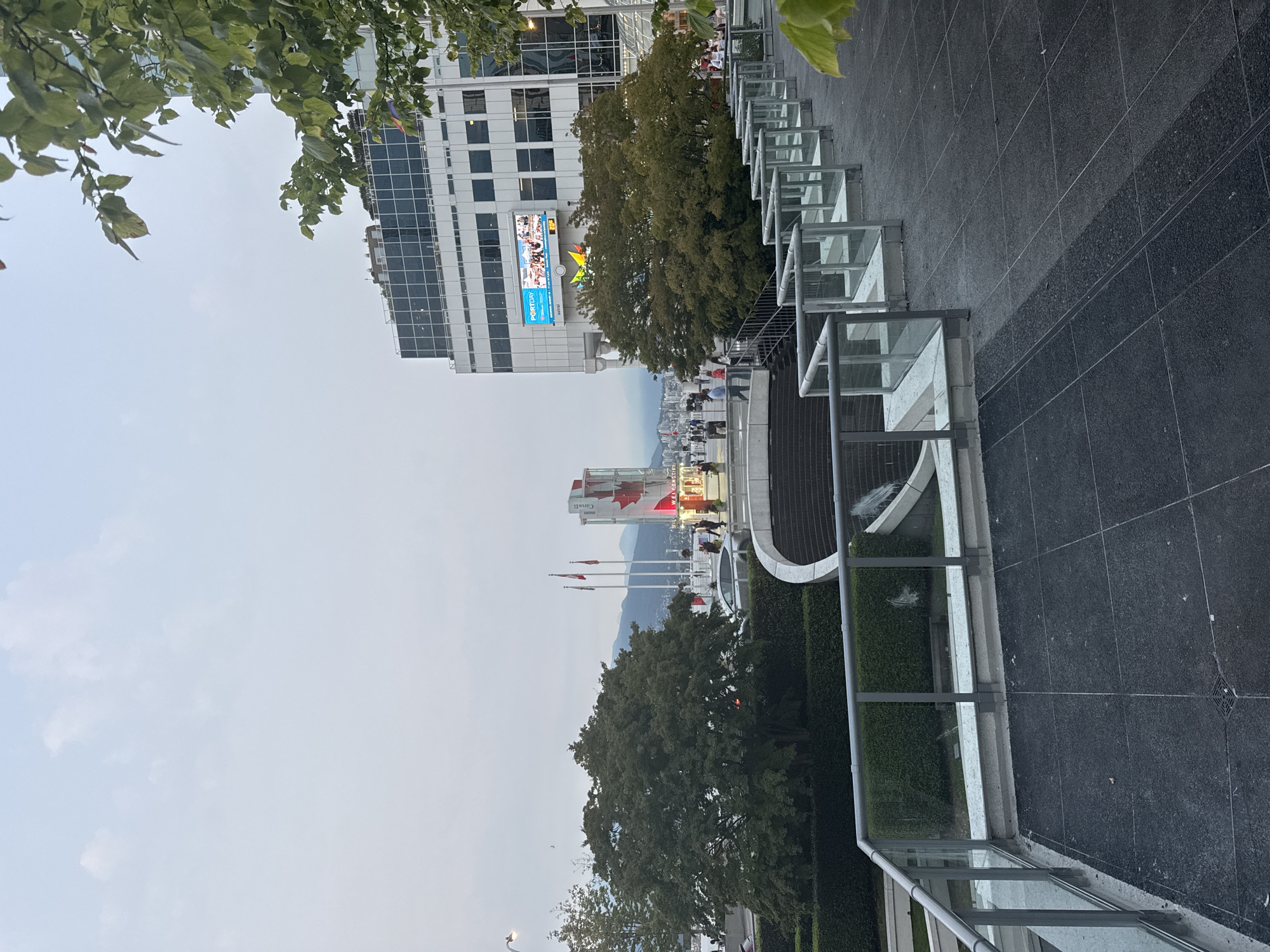 The image shows a view of the Canada Place building in Vancouver, Canada, from a raised vantage point. The Canada Place building, with its distinctive sails, is visible in the background. In front of the building, there is a large tree. A paved area with glass railings is in the foreground, with a set of stairs leading down. The sky is overcast. Several people are visible walking around the area. There is a large digital display screen on the side of the Canada Place building, showing an image of people on a boat. There are also Canadian flags flying. The overall scene appears to be a public plaza or promenade area.