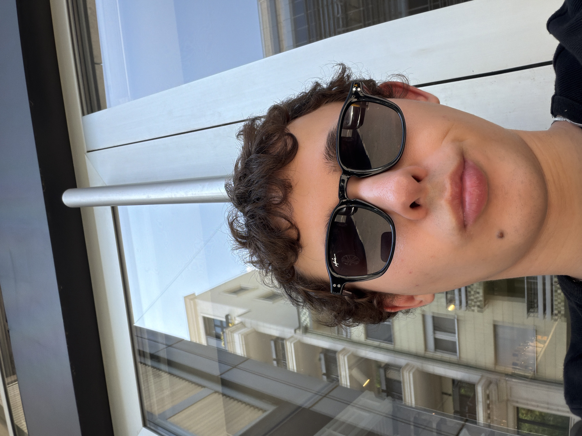 The image presents a close-up, chest-up selfie of a young man looking directly at the camera. He has dark, curly hair and is wearing black-framed sunglasses with dark lenses; a stylized 'M' logo is visible on the left temple. His lips are slightly pursed, and a small, dark mole is present on his lower right cheek. He wears a dark-colored garment, possibly a sweater or hoodie, and a silver chain is visible around his neck.
The man is positioned in front of a large window, which reflects the surrounding urban environment. To the left of the man, the window reflects a multi-story, light-colored building, characteristic of residential architecture in Vračar Urban Municipality, Serbia. This reflected building features multiple balconies, some with visible railings and decorative elements. The reflection also shows structural elements that suggest either internal building components or further reflections of other buildings. To the right of the man, a prominent vertical, light-gray, cylindrical structural element is visible, positioned against a background of reflections dominated by blue sky and less distinct building structures. Reflections within the man's sunglasses include a partial view of a vehicle, with what appears to be a headlight or grille in the right lens and a wheel or part of a car in the left lens.