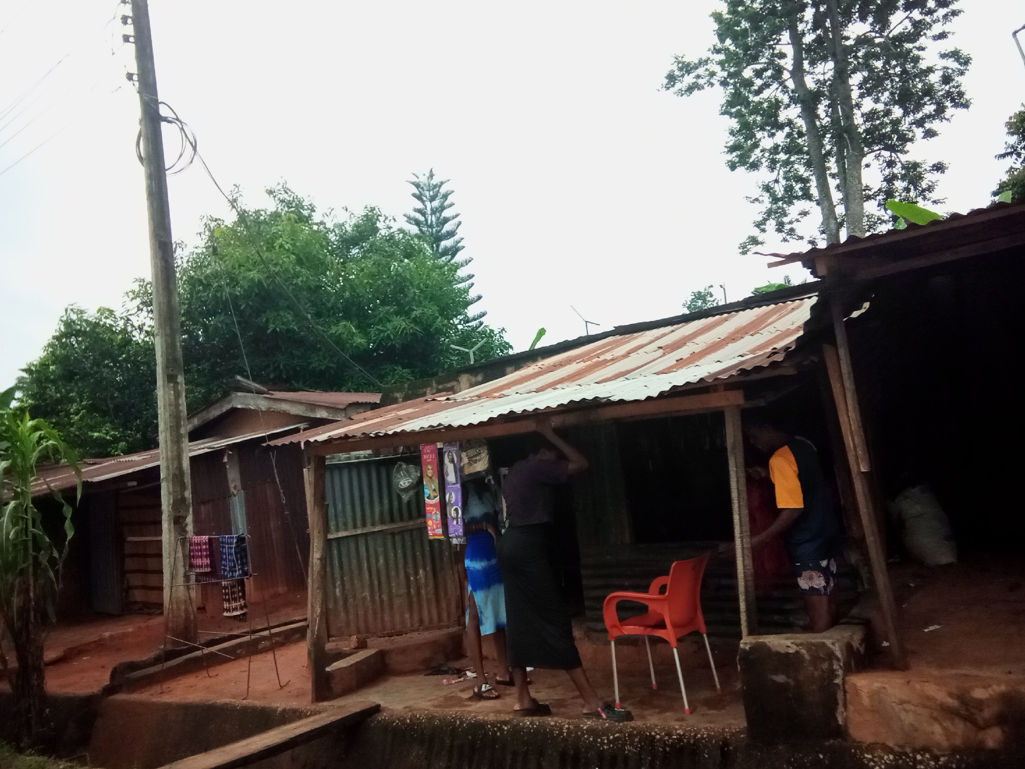 The image depicts a street scene in Oko Town, Nigeria, featuring a makeshift shop with a corrugated iron roof. Several individuals are visible, some interacting with the shop's contents, while others are standing nearby. An orange plastic chair is placed outside the shop. The environment appears to be an outdoor setting with some vegetation and trees in the background, under an overcast sky. The ground is reddish-brown, suggesting a dirt or clay surface. A utility pole with wires stands to the left of the frame, and various items, including textiles, are displayed at the shop.