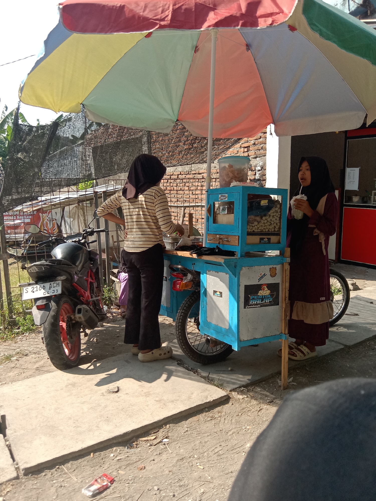 In Sugihwaras, Indonesia, a food cart vendor stands near a brick building and a fence, under a colorful umbrella. She wears a headscarf and a patterned dress. A customer, also wearing a headscarf and a striped shirt, is on the other side of the cart. The cart is blue and white, with a display of food. A motorcycle is parked beside the cart. The ground is a mixture of dirt and pavement.