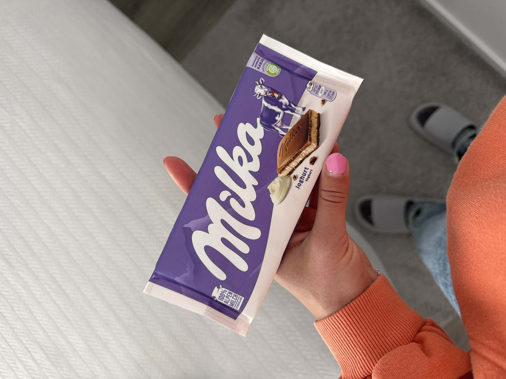 A person is holding a Milka Joghurt (Yoghurt) chocolate bar. The packaging is predominantly purple and white, with the Milka logo prominently displayed. The wrapper also features an image of the Milka cow. The hand holding the chocolate bar has a fingernail painted pink. In the background, there is a white textured surface (possibly a bedspread) and a glimpse of a person wearing light blue jeans and grey slippers. The location is not specified within the image. It is not possible to tell from the image if the location is in Blagoevgrad, Bulgaria.