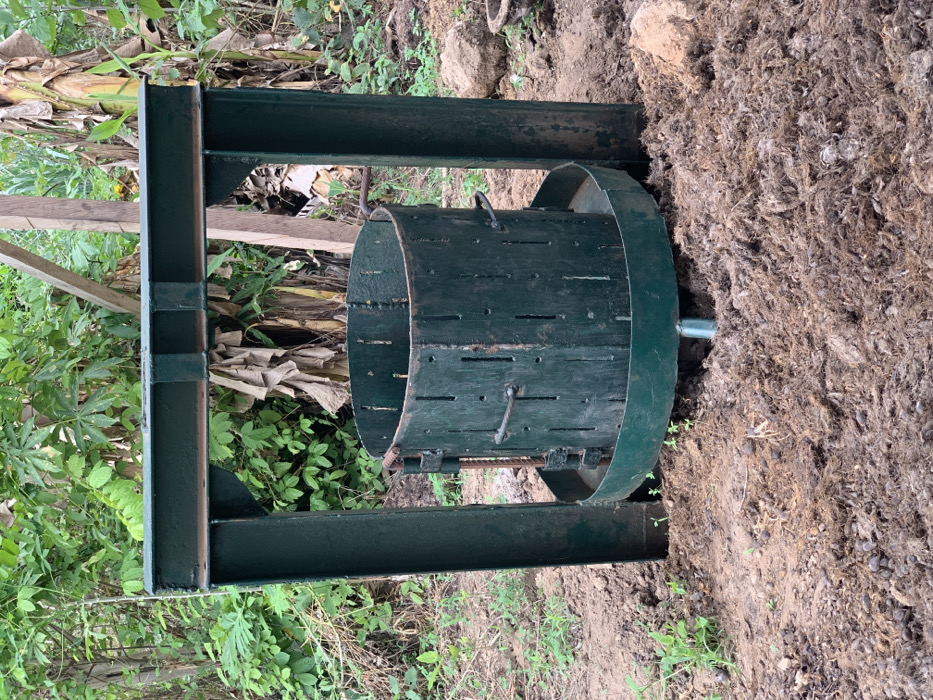 The image shows a large, green metal press, likely for extracting juice from fruit or other materials. The press is standing on a dirt ground that appears to be in a backyard or garden area. There is a dense patch of green foliage to the left of the press, possibly banana plants. The press is made of sturdy metal and appears to be in good condition. It features a round, perforated drum that rotates within a sturdy metal frame. The image appears to be taken on a sunny day, as there is no shadow cast by the press. The coordinates 7.119, 5.362 are located in the Egbeda area of Lagos, Nigeria.