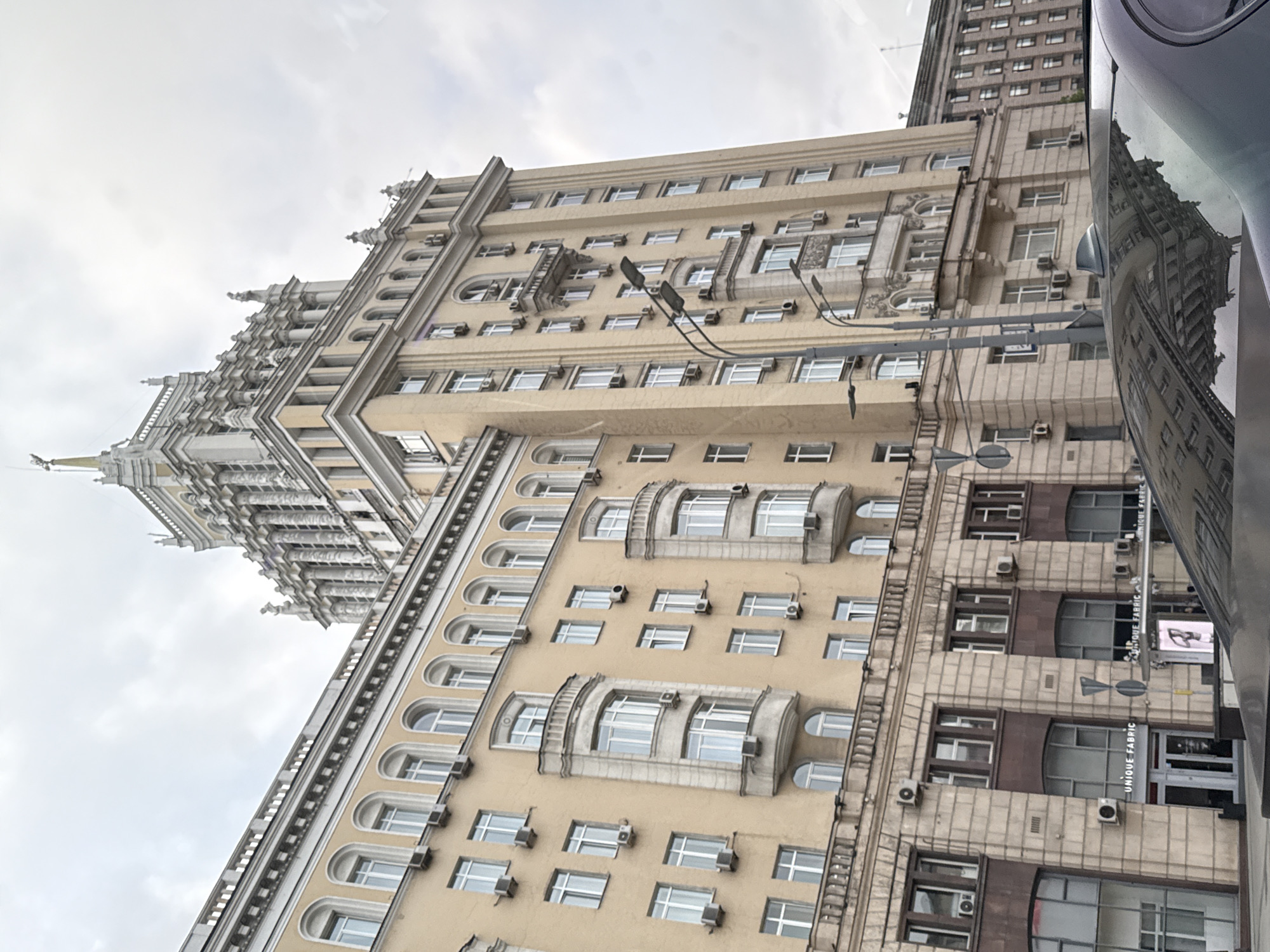 The image shows a building in Moscow, Russia. The building is tall and has many windows. It is made of stone or concrete. There are a few air conditioners visible on the building. In the reflection on the car, the building is visible from the bottom up.