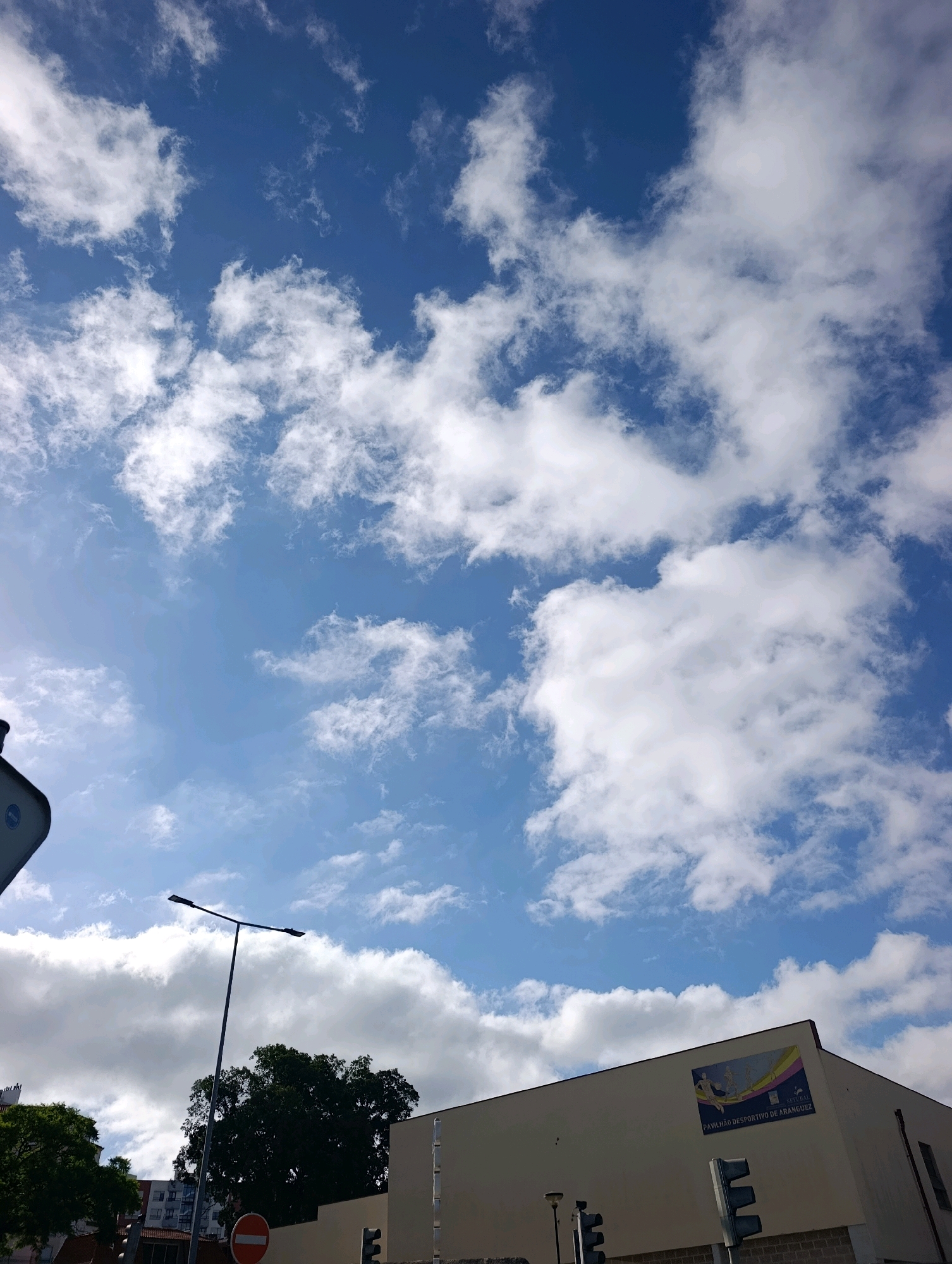 The image captures a view of the sky and buildings in Setúbal, Portugal. The sky is a bright blue with scattered white cumulus clouds. In the foreground, a portion of a light pole is visible, along with a traffic light and a circular sign with a red border and a white horizontal bar. To the right, a light-colored building stands, featuring a mural on its upper section. The mural depicts stylized figures in motion, with a banner below that reads "Pavilhão Desportivo de Aranguez". This indicates the building is likely the Aranguez Sports Pavilion. Trees and other buildings are also visible in the background, suggesting an urban or suburban setting. The overall scene is one of a sunny day in the city.