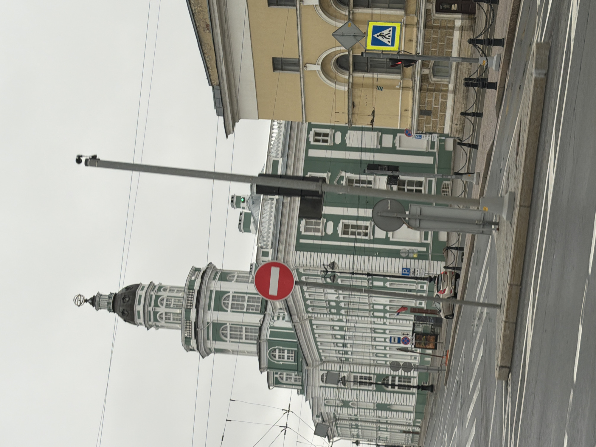 The image captures a street scene in Saint Petersburg, Russia, focusing on the Academy of Arts building and surrounding architecture. The foreground features a grey asphalt road with visible lane markings, including pedestrian crosswalks. A raised concrete curb lines the sidewalk.
In the mid-ground, several street elements are present. A tall, grey lamppost with a traffic camera is positioned on the right side of the road, angled towards the left. To the left of this lamppost, a red and white circular "no entry" traffic sign is visible on another pole. Adjacent to the "no entry" sign is a blue and white square sign depicting a pedestrian crossing. A traffic light, showing red, is mounted on a pole to the right of the pedestrian crossing sign. Further left, another lamppost with a dark pole and a street lamp is visible. A small, grey pedestrian signal pole stands between the lampposts. A silver car is parked on the right side of the road, behind a black metal chain-link fence.
The background is dominated by two prominent buildings. On the left is the Academy of Arts, a grand structure characterized by its green and white facade, numerous windows, and an ornate dome topped with an astronomical instrument. The building features classical architectural elements with white columns and decorative moldings. On the right is a yellow building with a light-colored cornice, featuring arched windows and a textured brick facade. A Russian flag is seen on a flagpole between the two buildings. Overhead, power lines crisscross the sky, which is overcast and a pale grey.