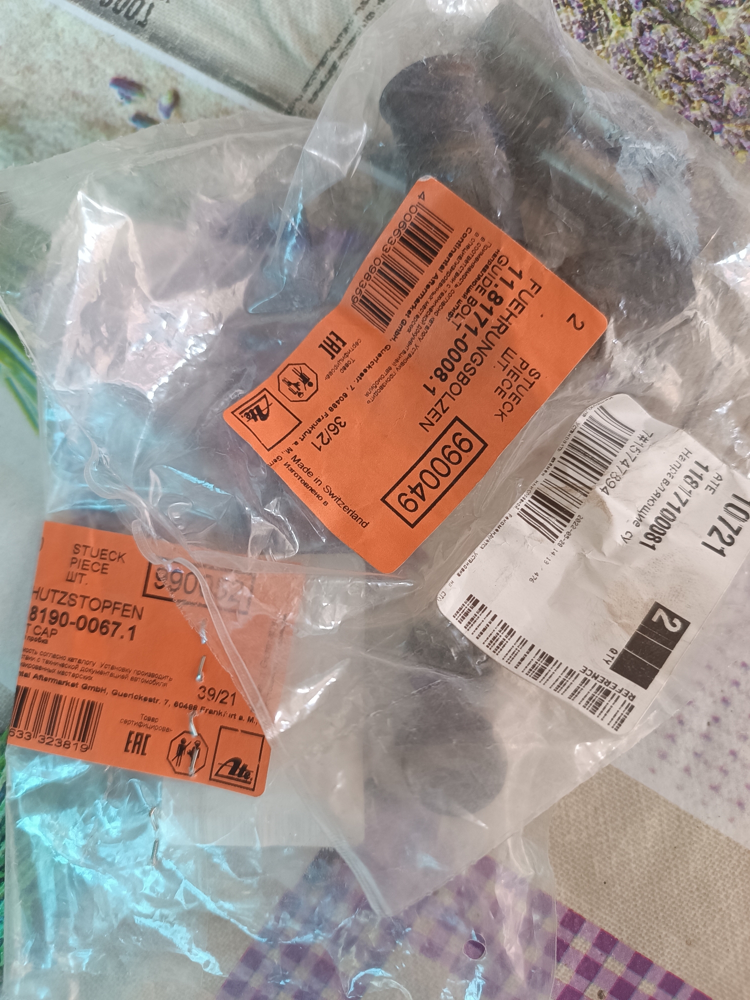 The image shows several plastic bags containing automotive parts. One bag has an orange label with the text "STUECK", "PIECE", "ШТ.", "HUTZSTOPFEN", "8190-0067.1", "T CAP", and a barcode. Another orange label, partially visible, has the text "36/21" and "Made in Switzerland". A third label, also orange, has the text "FUEHRUNGSBOLZEN", "11.8171-0008.1", "STUECK", "PIECE", "ШТ.", and a barcode. A white label has the text "10721" and "11877100081". The image is a top-down view of these items lying on a textured surface. There is no indication of the city of Vypasne, Ukraine, in the image.