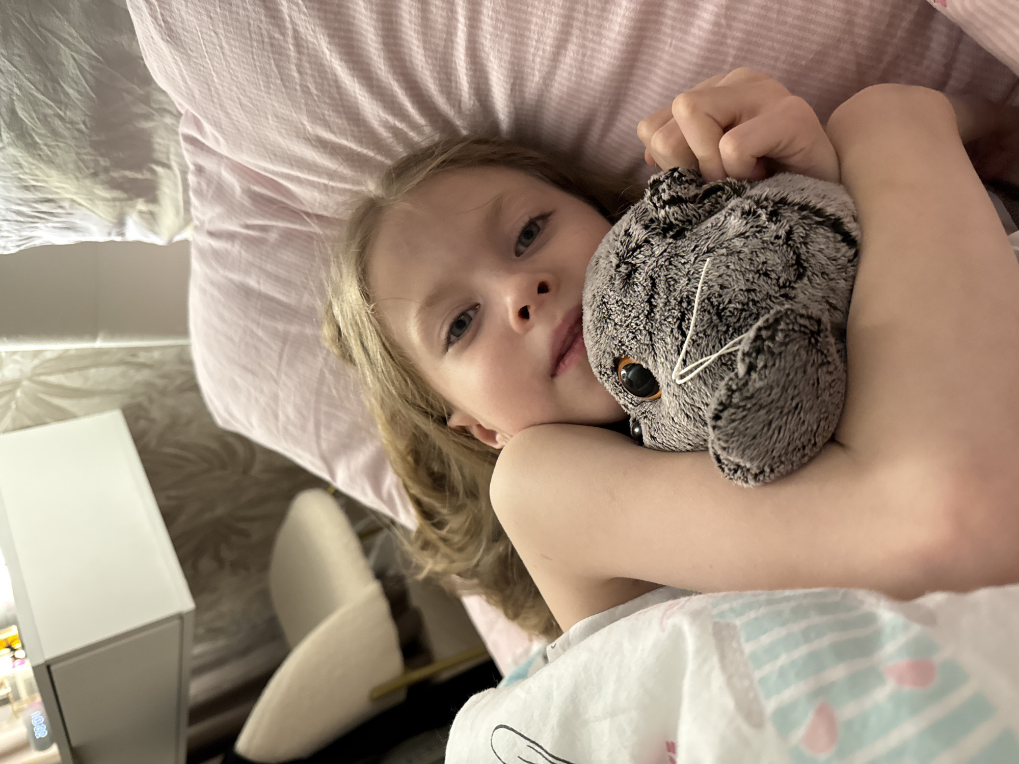 A young, light-skinned female child is lying in a bed. She is cradling a grey, plush owl toy. The bed has pink striped pillowcases and a white patterned blanket. A white bedside table is visible in the background. There is no information to indicate the location is Izhevsk, Russia.