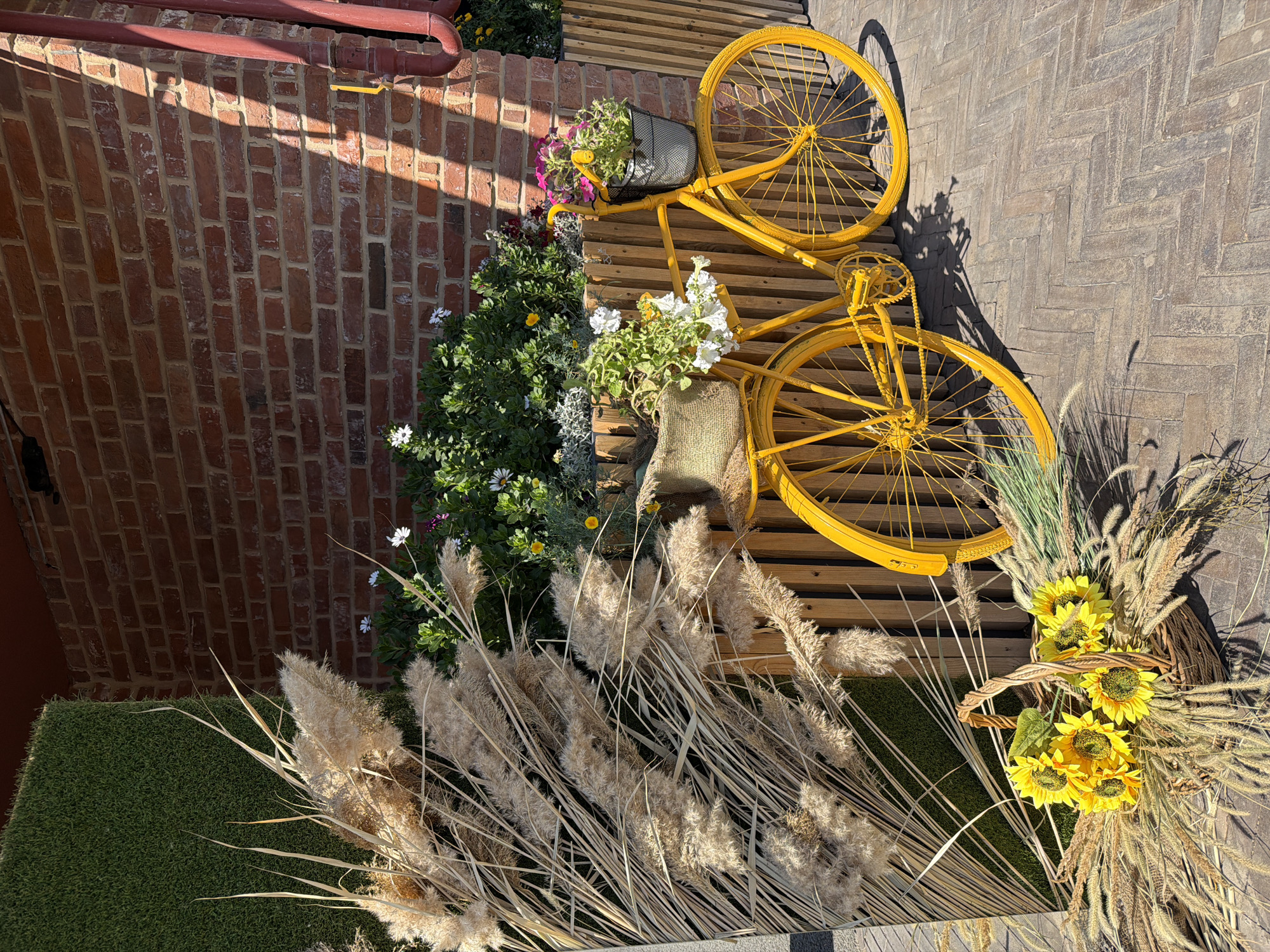 In Buzuluk, Russia, a yellow bicycle is prominently displayed as a decorative element against a textured brick wall and a backdrop of artificial green turf. The bicycle is adorned with potted flowers in its front basket and a woven sack on its rear rack, suggesting a floral or autumnal theme. Surrounding the bicycle, various dried grasses and a basket of sunflowers add to the rustic and festive atmosphere. The scene is illuminated by natural sunlight, casting shadows that highlight the textures of the brick wall and the surrounding vegetation. The ground is paved with herringbone patterned bricks.