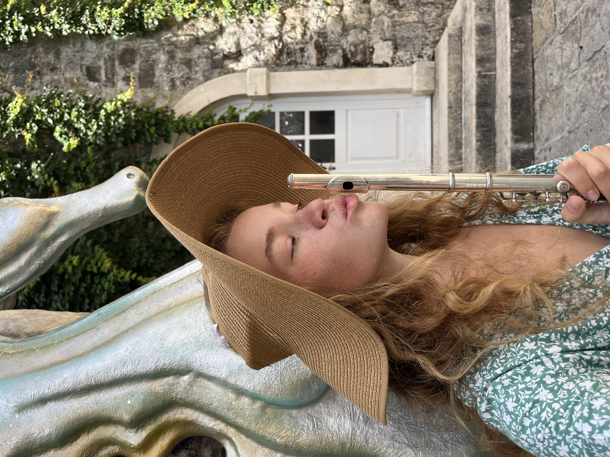 An outdoor, brightly lit image captures a young person lying down, holding a silver flute to their lips. The person, appearing female, has long, wavy, reddish-blonde hair and light skin with visible freckles. They wear a wide-brimmed straw hat and a green dress adorned with a white floral pattern. Their eyes are mostly closed, suggesting a moment of relaxation, concentration, or repose.
The background features a rustic stone wall, partially covered in vibrant green ivy or climbing plants. Centered behind the person's head is a white wooden door with a distinctive multi-paned window. To the right, light-colored stone steps or vertical architectural elements are visible. The person is resting on a light, textured, and slightly sculptural surface, possibly a large statue, bench, or decorative architectural element. The bright illumination indicates a sunny day. The scene evokes a tranquil setting, consistent with the historical and scenic character of Budva, Montenegro. There is no visible text in the image.