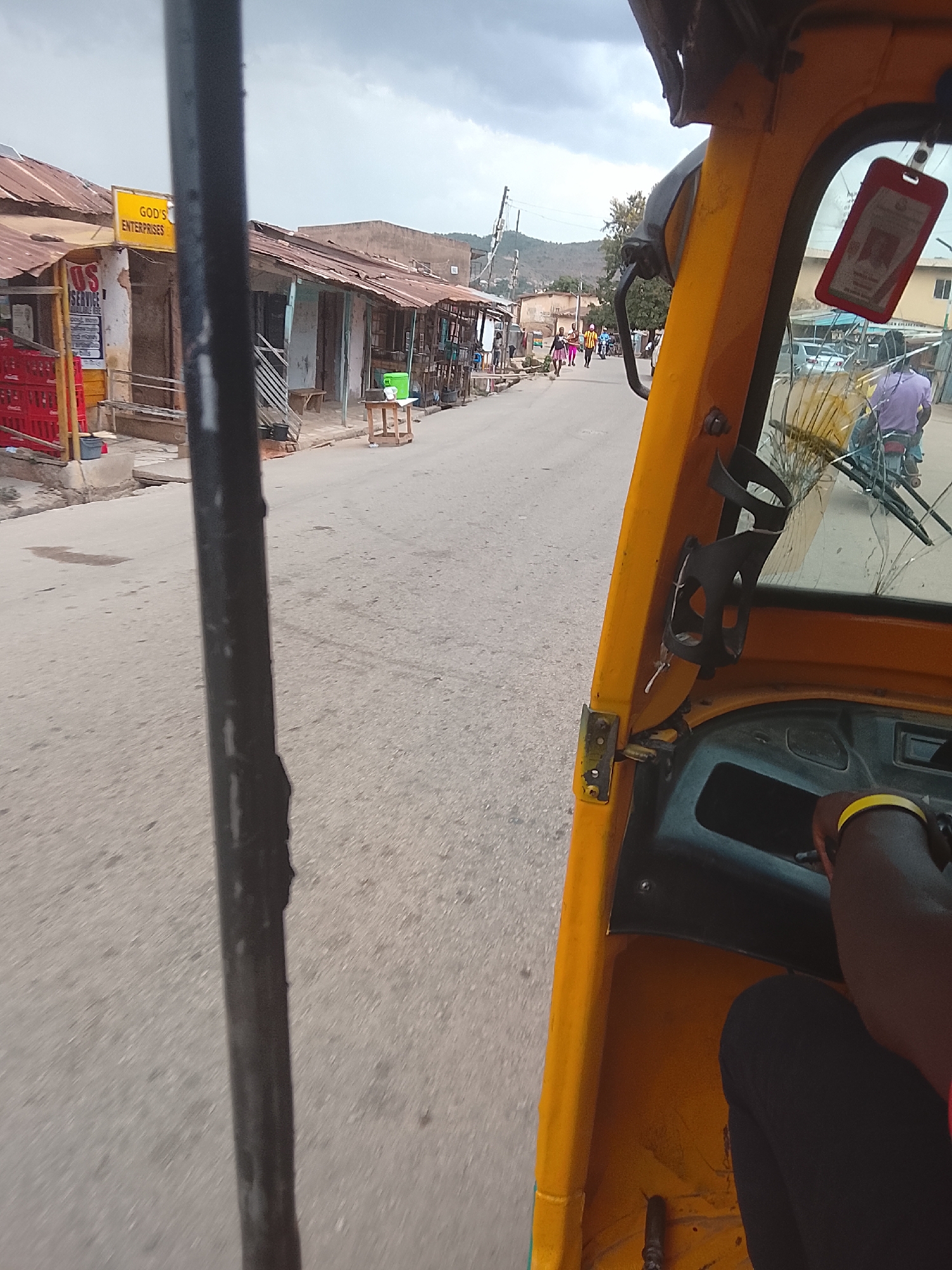 The image shows a view from inside a yellow auto-rickshaw (keke) traveling on a street in Jos, Nigeria. The street is lined with single-story buildings with corrugated iron roofs. Some of the buildings appear to be small businesses, indicated by signage (e.g., "God's Enterprises"). Pedestrians are visible further down the street. The keke has a cracked windshield. The driver's arm, wearing a yellow wristband, is visible in the foreground. The sky is overcast.