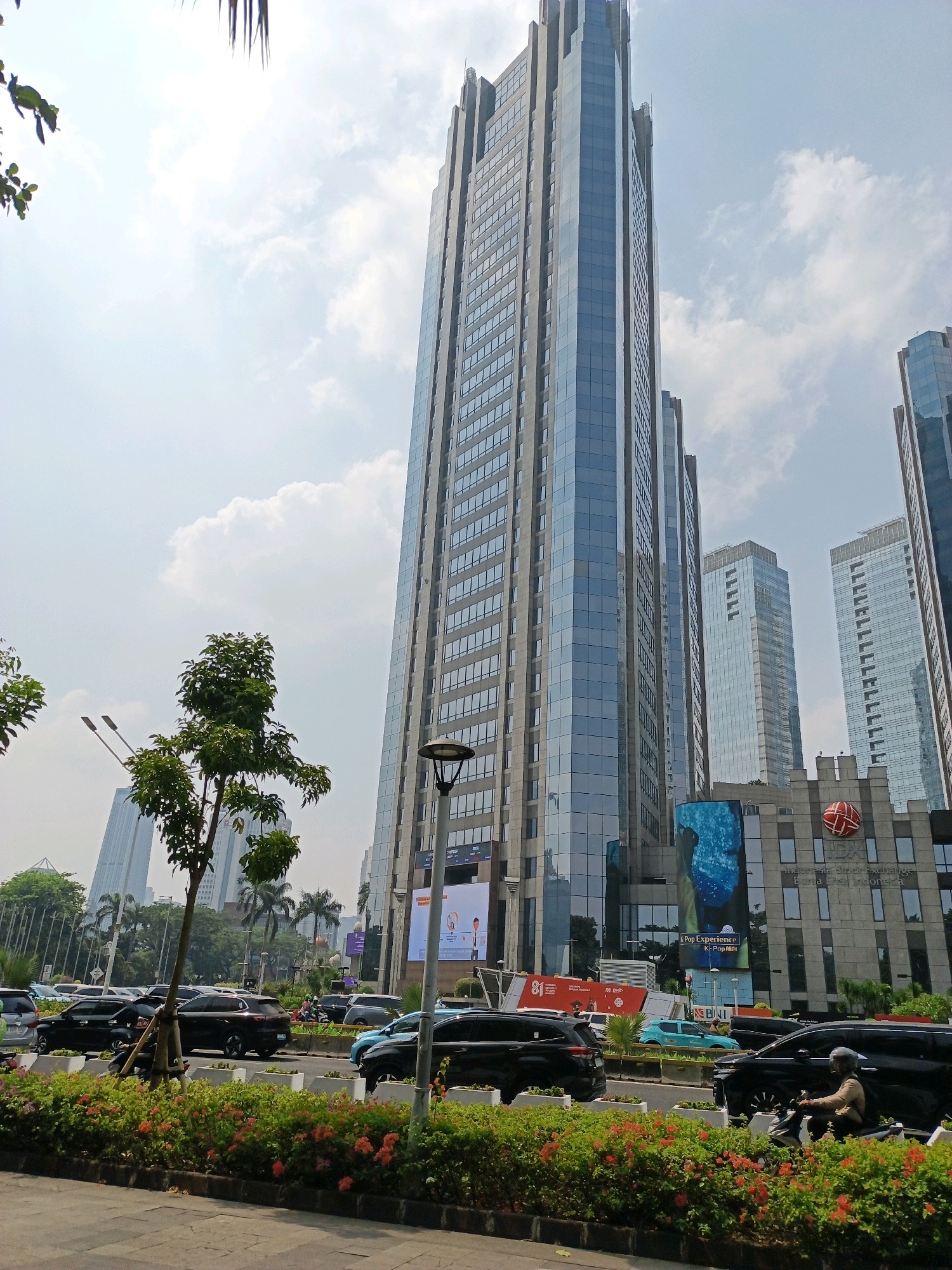 The image captures a street scene in Central Jakarta, Indonesia, dominated by a towering, modern skyscraper with a glass facade. The skyscraper is flanked by other, slightly shorter glass-fronted buildings, indicative of a central business district. In the foreground, a lush median strip is covered with vibrant red and orange flowers, bordered by a concrete sidewalk. Cars, including black SUVs and sedans, are visible on the multi-lane road, suggesting moderate traffic. A person rides a motorcycle in the right lane, wearing a helmet and a light-colored jacket. Several trees, including a prominent leafy tree in the middle ground, add greenery to the urban landscape. The sky is partly cloudy, with bright daylight illuminating the scene. A street lamp with a modern design stands near the leafy tree. Billboards and digital displays are present on the lower levels of the buildings, advertising various products and services. The overall impression is of a bustling, contemporary urban environment in Indonesia's capital city.