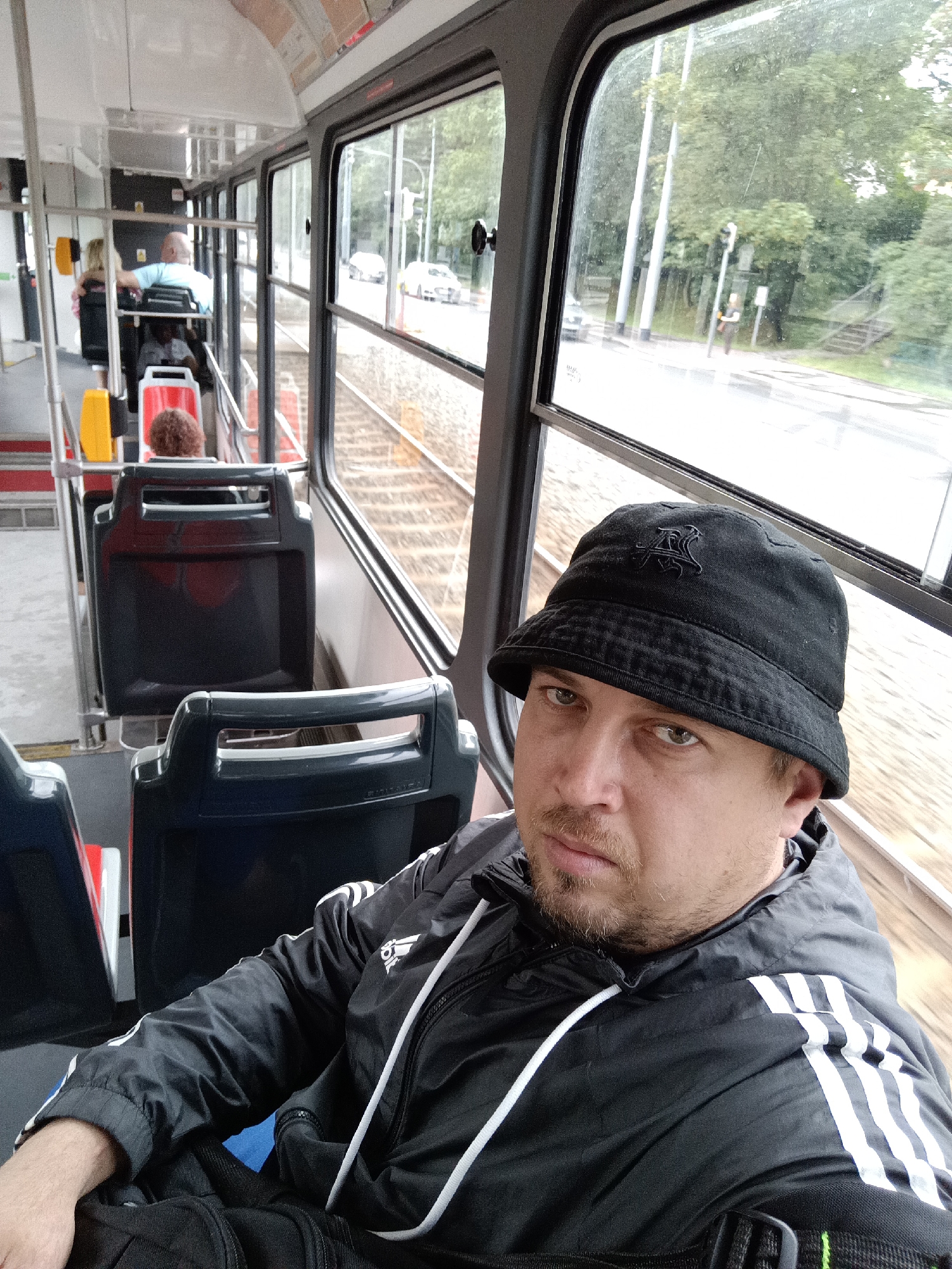 A man is sitting on a tram in Prague, Czechia. He is wearing a black bucket hat with an embroidered logo and a black Adidas jacket with white stripes. He is looking at the camera with a neutral expression. The tram interior is visible, with rows of dark seats and windows. Outside the window, trees and a street are visible, suggesting an urban setting. The weather appears overcast, and the tram is moving along tracks. Other passengers are visible in the background, some seated and others standing. One passenger in the foreground is seen from behind, with curly brown hair. The overall atmosphere is mundane, depicting a typical commute.