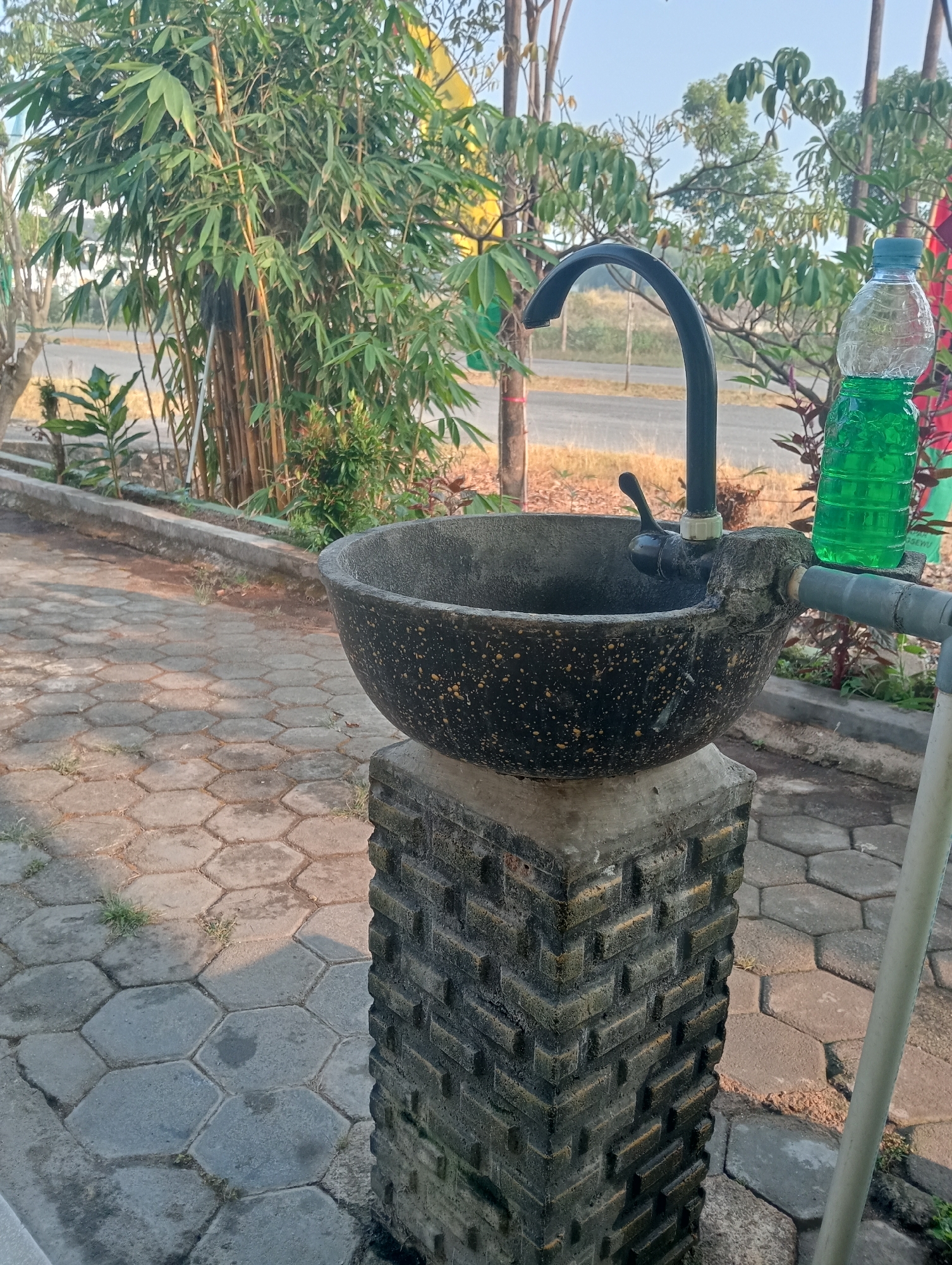 The image depicts an outdoor sink with a concrete basin and a decorative concrete pedestal. The sink features a black faucet with a lever handle. To the right of the sink, a green liquid, likely soap or disinfectant, is contained within a clear plastic bottle. The sink is situated outdoors on a paved area composed of hexagonal concrete tiles, some of which are moss-covered or damp. Behind the sink, there are various plants, including bamboo stalks and leafy shrubs. A road is visible in the background, beyond which are more trees and vegetation. The scene appears to be in a tropical or subtropical climate, given the type of foliage. The lighting suggests it is daytime, with shadows cast on the tiled ground. The location is identified as Sidoharjo, Indonesia. There are no people visible in the image.