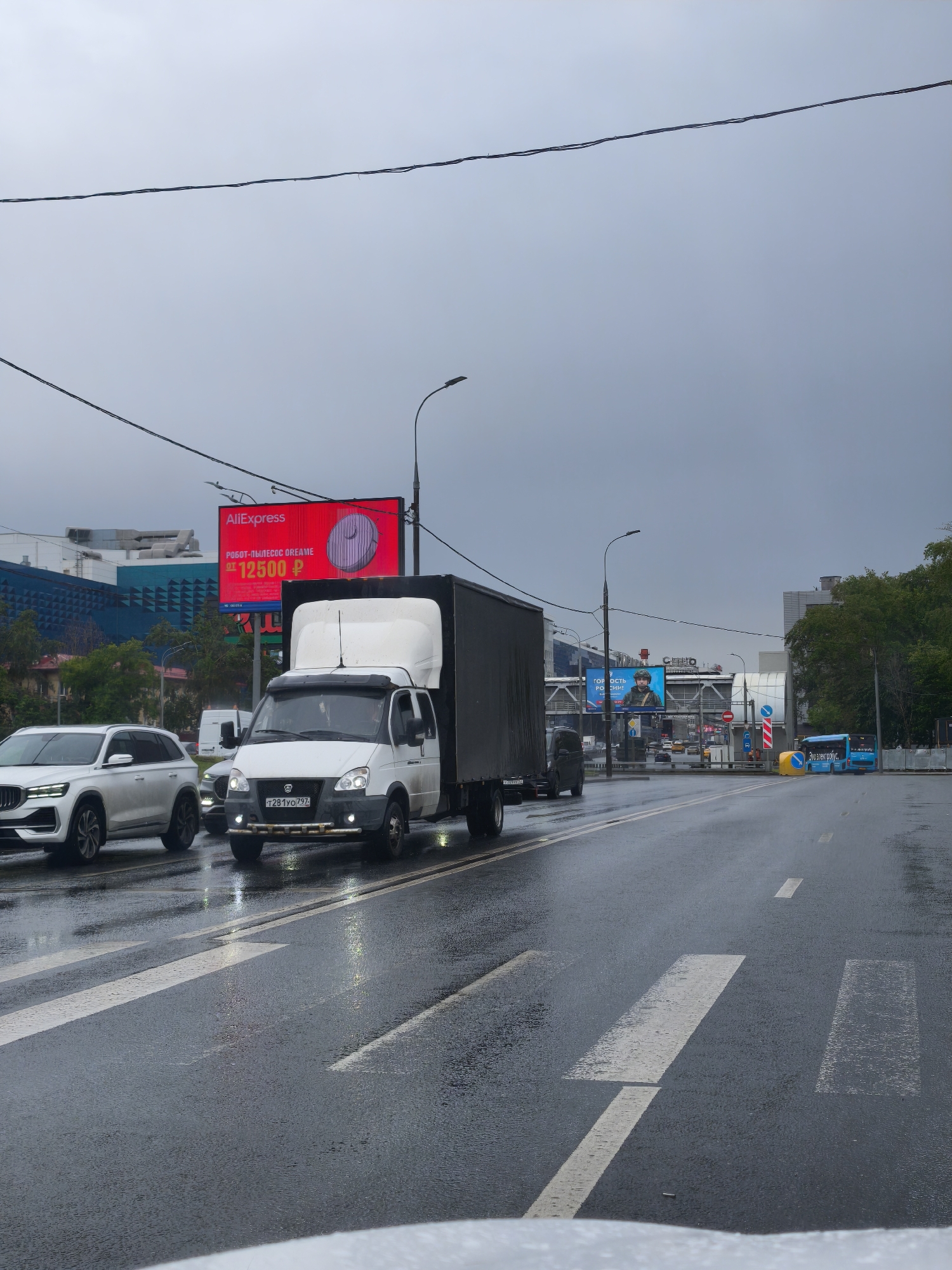 The image depicts a street scene in Moscow, Russia on a rainy day. The road is wet and reflects the ambient light. Several vehicles are present, including a white SUV, a white and black box truck with the license plate "T281YO797", and a blue bus further down the road. In the background, there are various buildings and billboards, one advertising AliExpress with a price of 12500 ₽. Overhead power lines are visible against the overcast sky. The road markings indicate a crosswalk.
