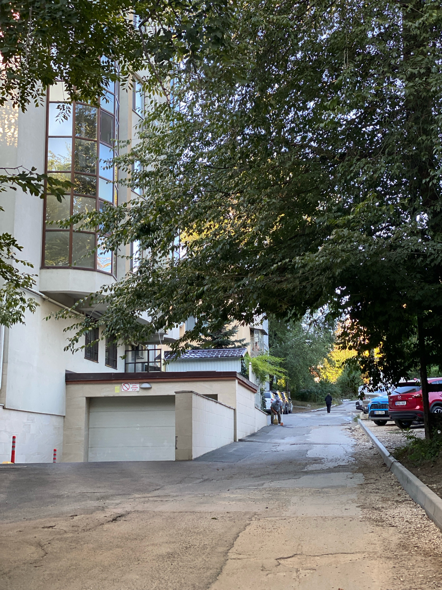 This image depicts a street scene in Chișinău, Moldova, featuring a modern building with large windows on the left, partially obscured by lush green trees. A driveway leads from the foreground to a garage door entrance under the building. Several cars are parked along the right side of the street, which slopes upwards. A person is visible walking up the street in the distance. A man is sweeping the pavement near a row of parked cars on the left. The road surface shows signs of wear and tear with visible cracks and patches. The scene appears to be set in a residential or mixed-use urban area during daylight.