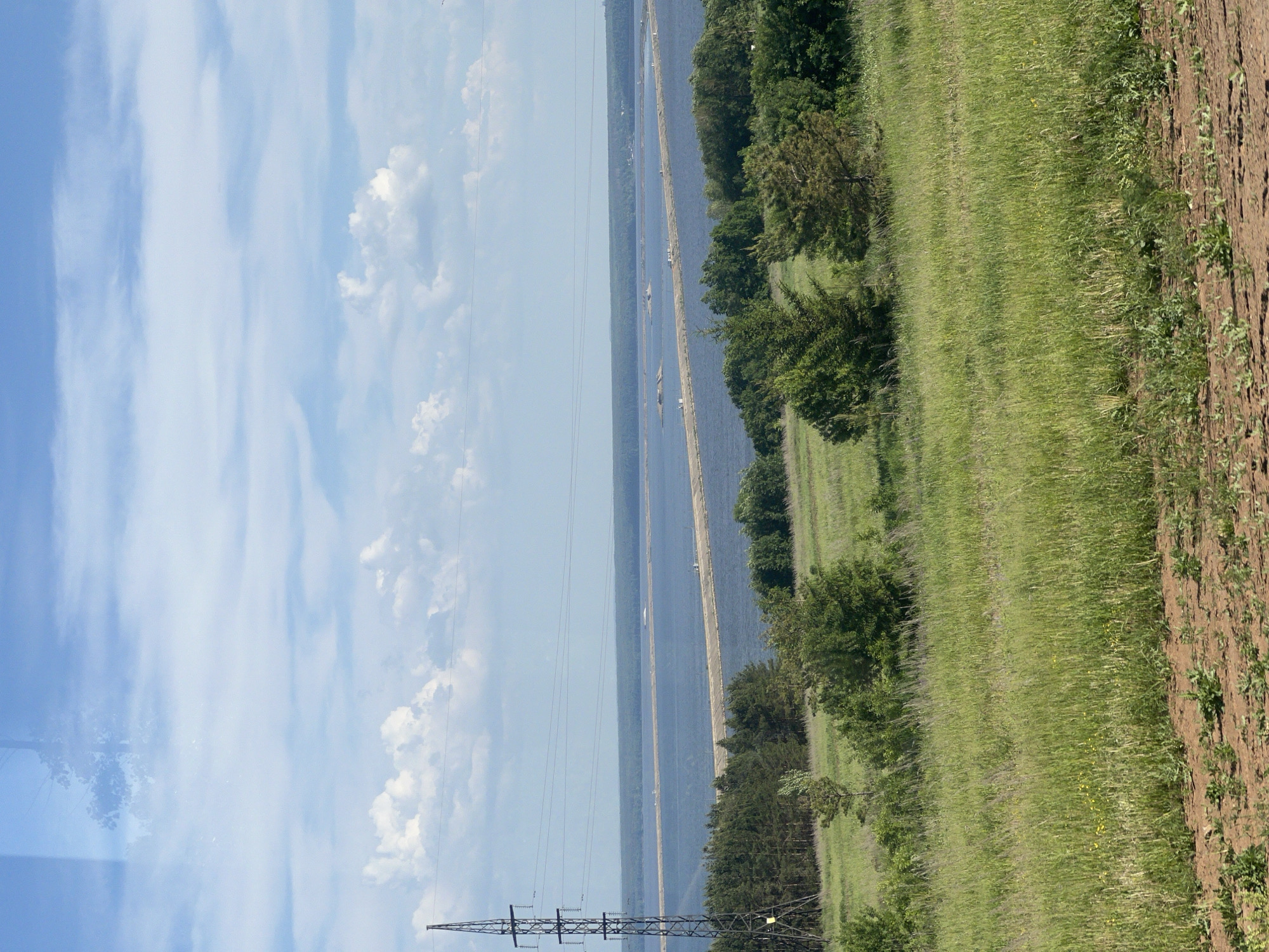The scene is a rural landscape in Белоус, Russia, featuring a river with two narrow, sandy islands running parallel within it. A high-voltage transmission tower is visible in the lower left corner of the frame. To the right of the river are patches of trees and green grassy fields, extending to a section of tilled soil or a dirt road. The sky occupies the left side of the image and is blue with streaks of white clouds.