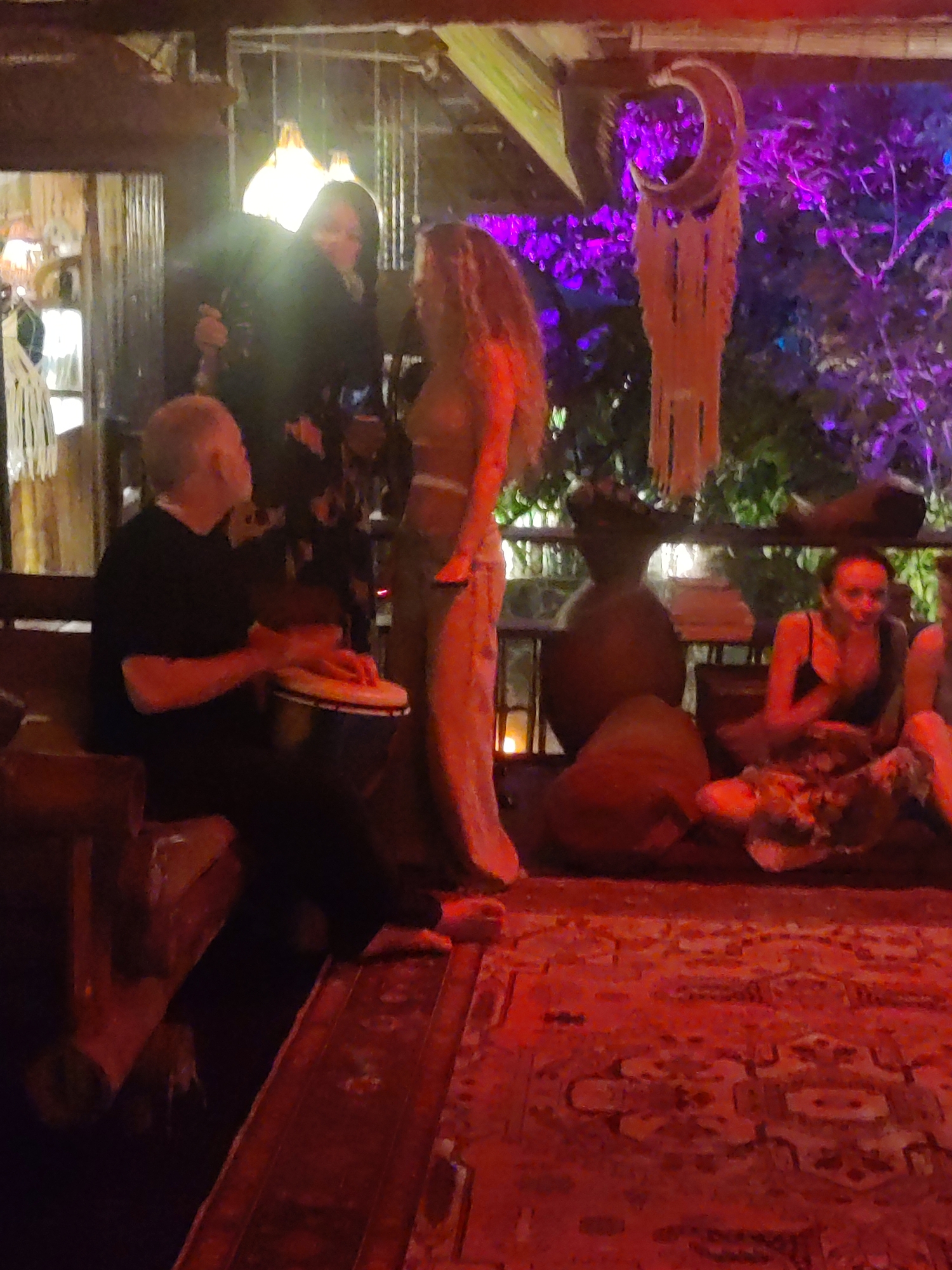 The image depicts an outdoor gathering, possibly at a lounge or restaurant in Lokaserena, Indonesia. A man seated on a wooden bench is playing a djembe. He is wearing a black shirt and dark pants. Two women are standing near the man playing the drum; one wears a dark patterned dress, and the other wears tan pants and a matching top. Two women sit on cushions near a large urn, one wearing a black tank top and patterned skirt. The scene is illuminated by both indoor and outdoor lighting, including string lights. A carpet covers the ground and light fixtures. The setting includes foliage and possibly decorative elements.