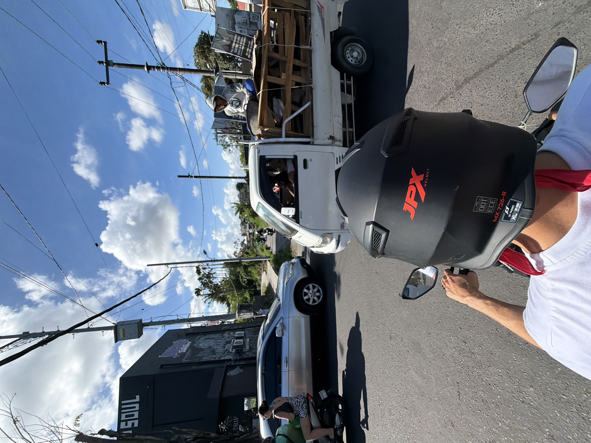 The image depicts a street scene in North Kuta, Indonesia. The perspective is from a motorcycle rider wearing a black "JPX" brand helmet with a red bandana. Other vehicles on the road include a white Kei truck carrying a person and wooden materials, and a silver SUV. A person is seen kneeling by a scooter. On the left is a black building with the word "SOUL" visible. Utility poles and power lines are also present. The sky is blue with scattered white clouds.