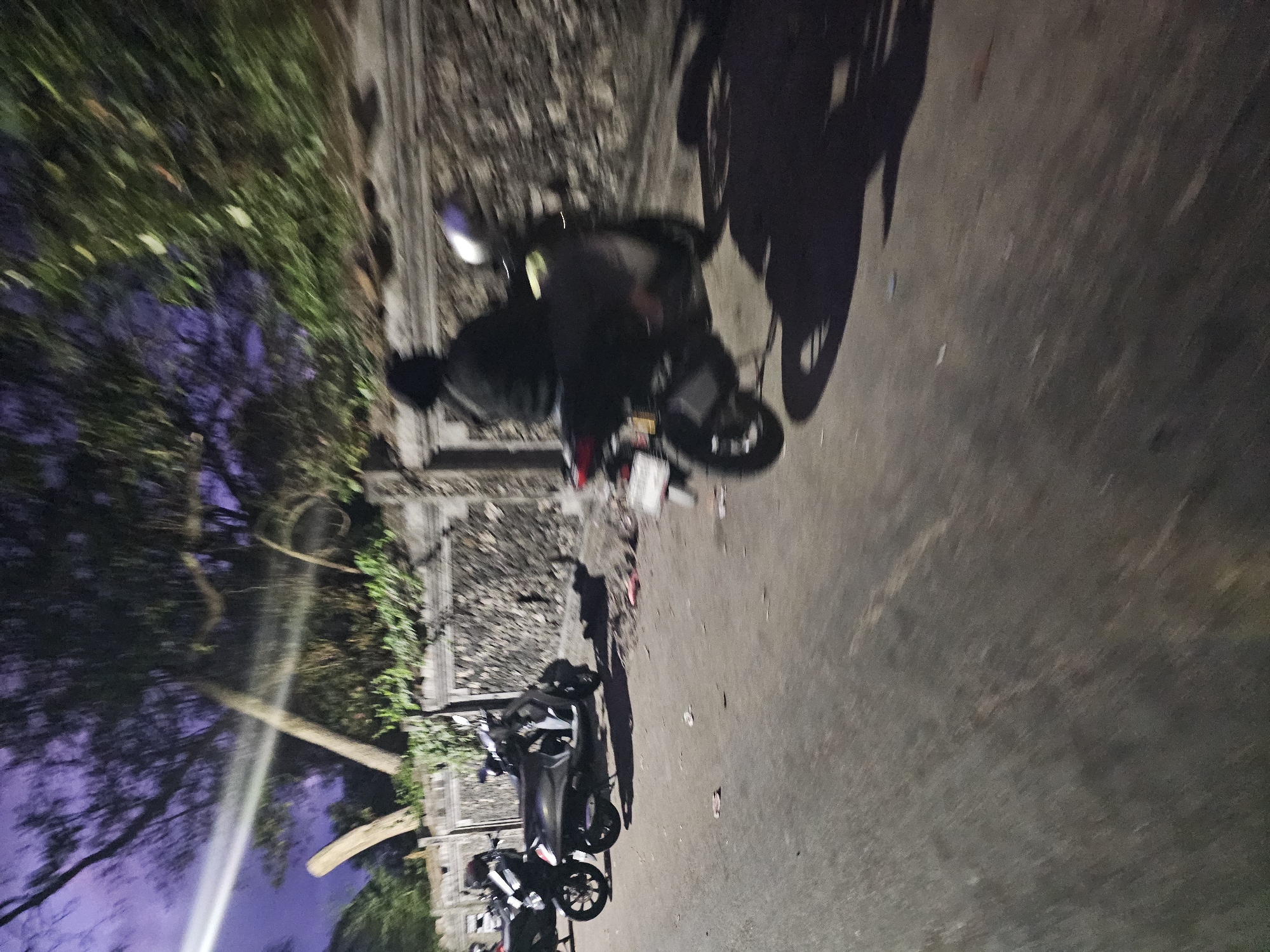 The image depicts a blurred nighttime scene on a street in Denpasar, Indonesia. A person is riding a scooter, their figure and the scooter appearing streaked due to motion. Several other parked scooters are visible on the left side of the street. The background features a stone wall and trees, with the sky showing a deep purple hue, indicative of twilight or night. The street itself is paved and appears somewhat uneven.