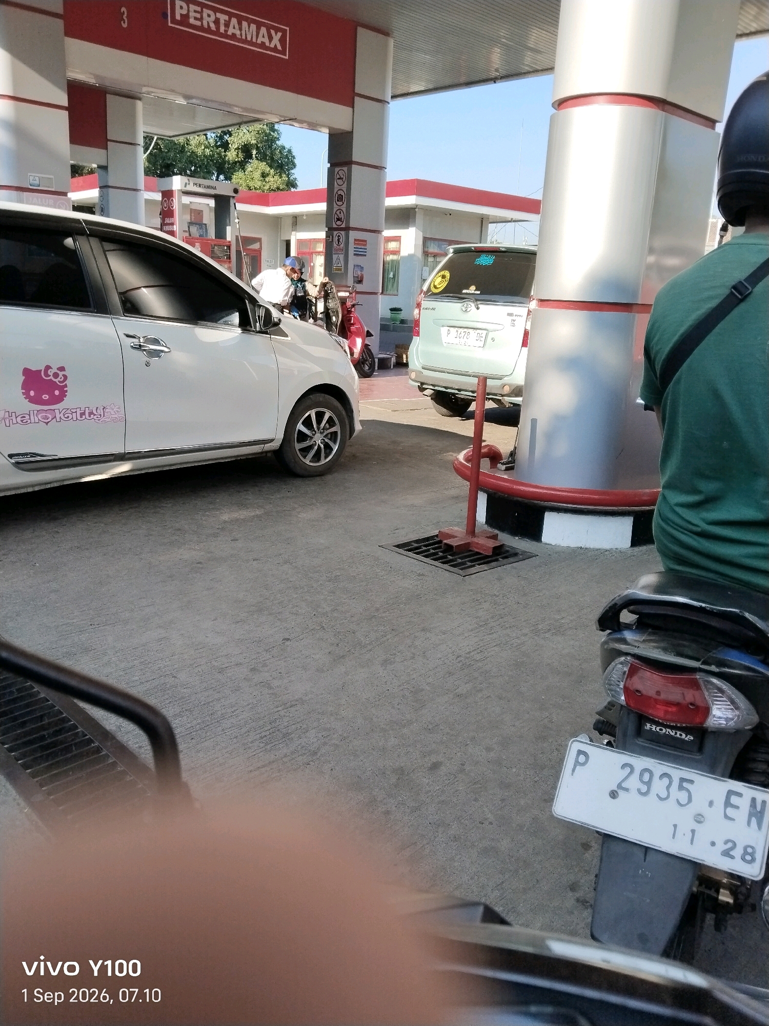 A rear-view shot captures a gas station in Situbondo, Indonesia, with a Pertamax sign visible above. In the foreground, the rear of a motorcycle with license plate P 2935 EN is parked on a paved surface. To the left, a white car adorned with a Hello Kitty sticker is parked at a gas pump. A person in a green shirt is seated on the motorcycle. In the background, a light-colored car is parked, and a red scooter is visible. The environment appears to be outdoors under a clear sky.