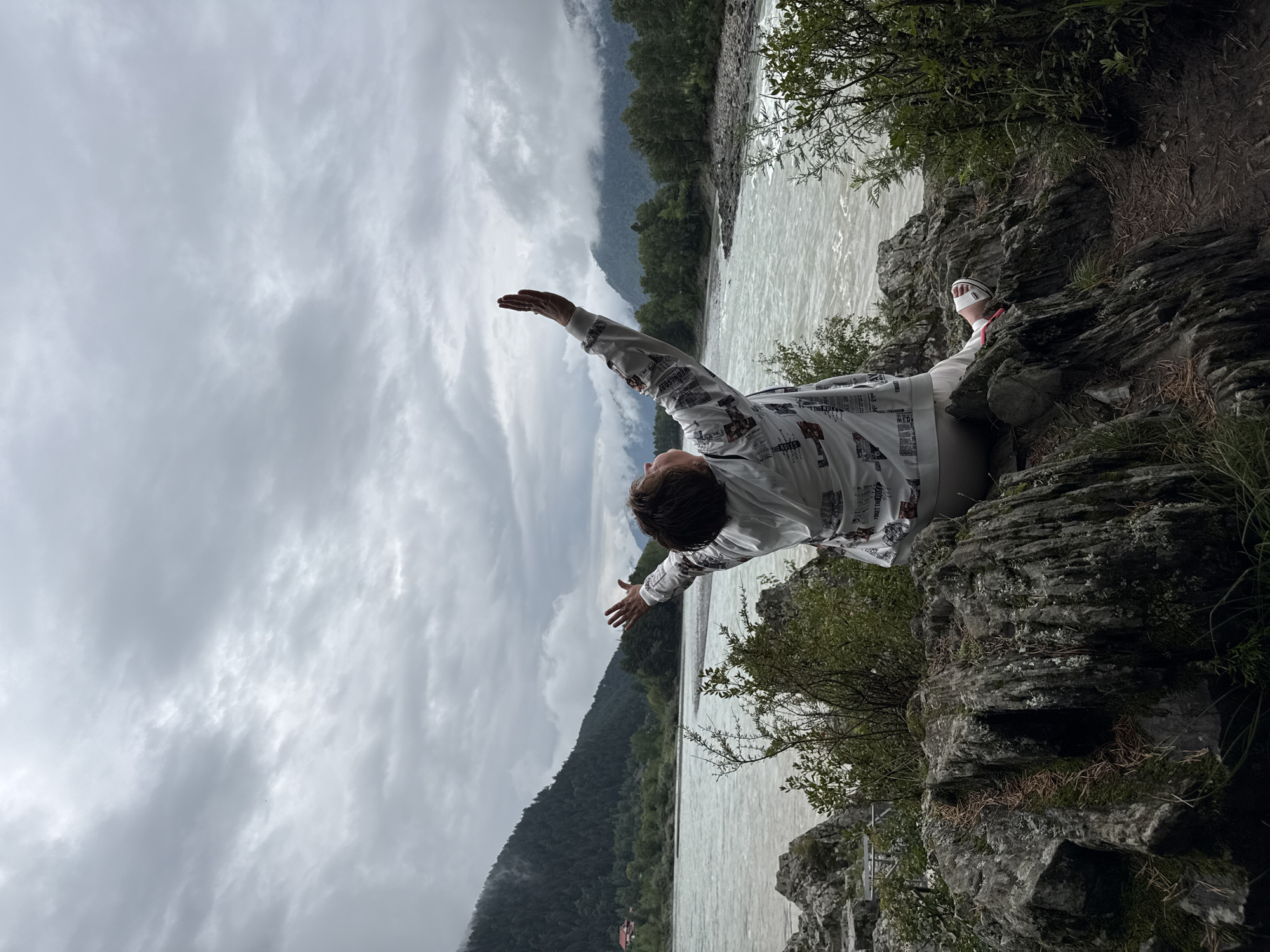A person is seated on rocks by a river with their arms raised to the sky. The person is wearing a white jacket with a graphic print and white pants. They are also wearing white sandals. The river is wide and appears to be flowing quickly. The background shows a mountainous landscape with trees and a cloudy sky. The location is the city of Elekmonar, Russia.