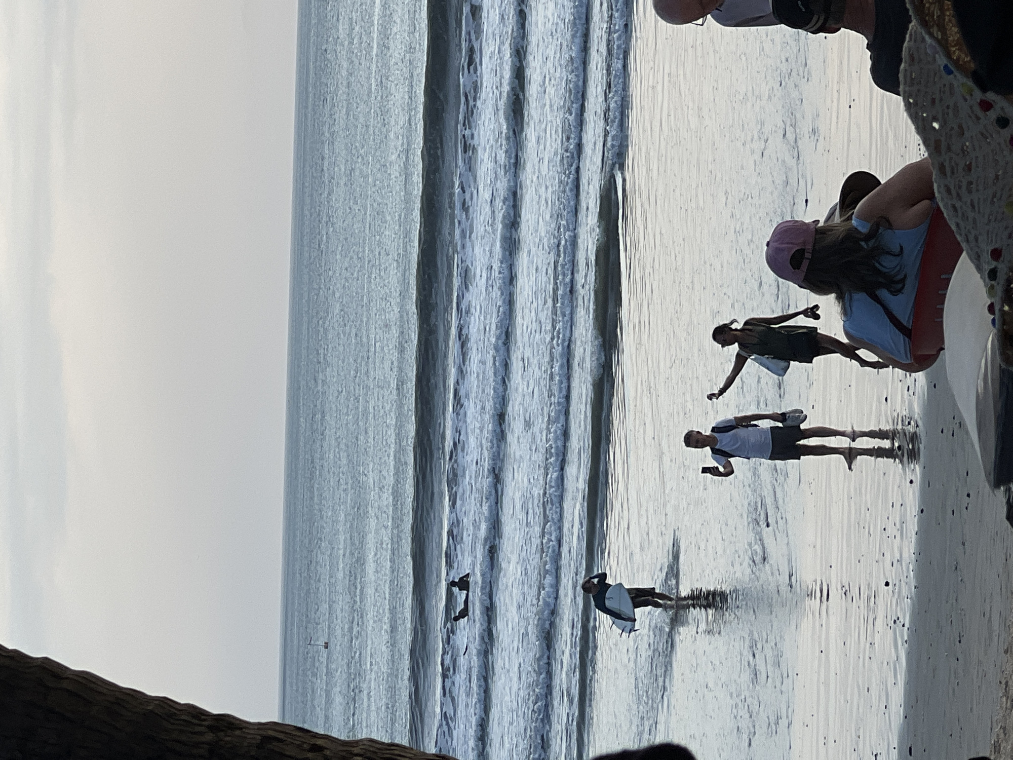 In Kuta, Indonesia, a group of individuals are present at a beach during what appears to be either early morning or late evening. The sea exhibits moderate waves, and several surfers are visible in the water. On the wet sand, a person is walking out of the water carrying a surfboard. Further inland, a man is holding up a mobile phone, possibly taking a photograph, while another person, a woman, is walking with her arm extended. In the foreground, two individuals are seated, one wearing a pink cap. The scene is framed by a large tree trunk on the left side of the image.