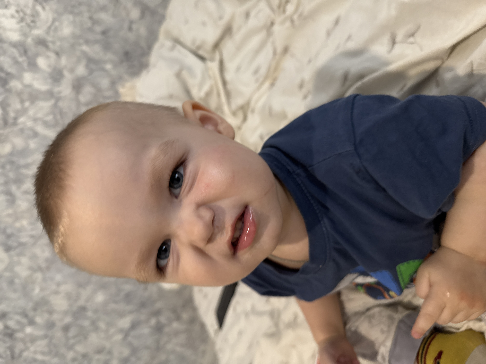 A close-up, high-angle shot shows a baby lying on a bed or soft surface, looking directly at the camera. The baby has fair skin, light brown hair, and striking blue eyes. A faint reddish mark is visible on the baby's left cheek. Their lips are slightly parted, revealing a few bottom teeth, and appear wet, suggesting drooling or recent feeding. The baby is wearing a dark blue short-sleeved t-shirt with hints of green and blue patterns at the bottom, suggesting a design or a colorful garment underneath like a diaper or shorts.
The background consists of two distinct textures: on the left, a gray and white fluffy or textured blanket/rug, and on the right, a lighter, off-white quilted or patterned blanket. The overall lighting suggests an indoor setting, likely a home, during the day. The baby's expression is one of curiosity and engagement, possibly a slight smile or a playful gaze. No text is visible in the image.