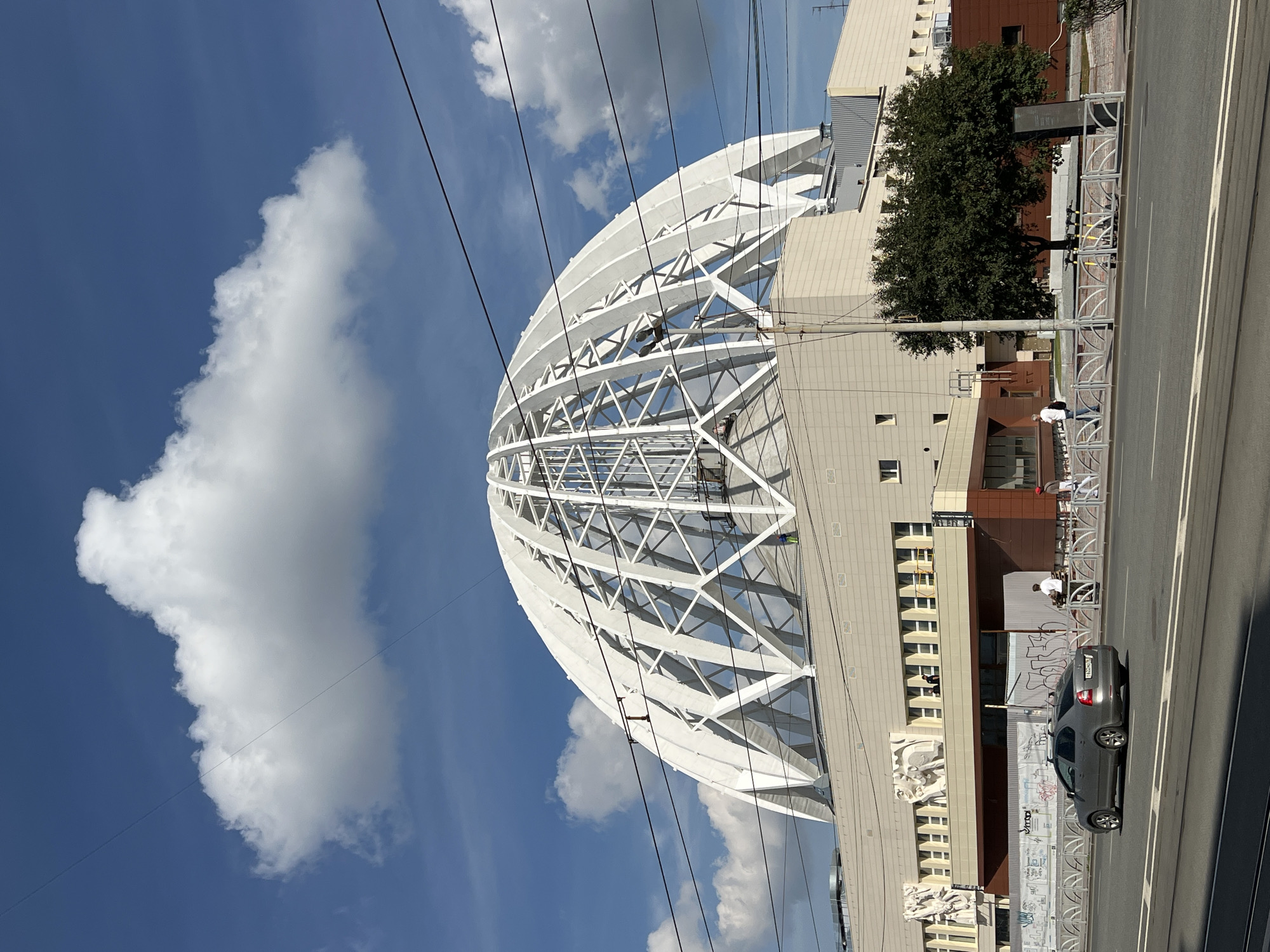 The image shows a view of the Yekaterinburg Central Stadium (now Ekaterinburg Arena) in Yekaterinburg, Russia. The stadium features a large, white, ribbed dome structure supported by a geometric framework, which is partially visible against a bright blue sky with scattered white clouds. The lower part of the building is a light-colored, rectangular structure with rows of windows. Relief sculptures are visible above the windows on the left side of the building. In the foreground, a multi-lane road runs horizontally across the image. A silver car is driving on the road, and several pedestrians are walking on the sidewalk. Overhead power lines are strung across the upper portion of the frame, and a green tree stands to the right of the building. The scene appears to be set on a sunny day.
