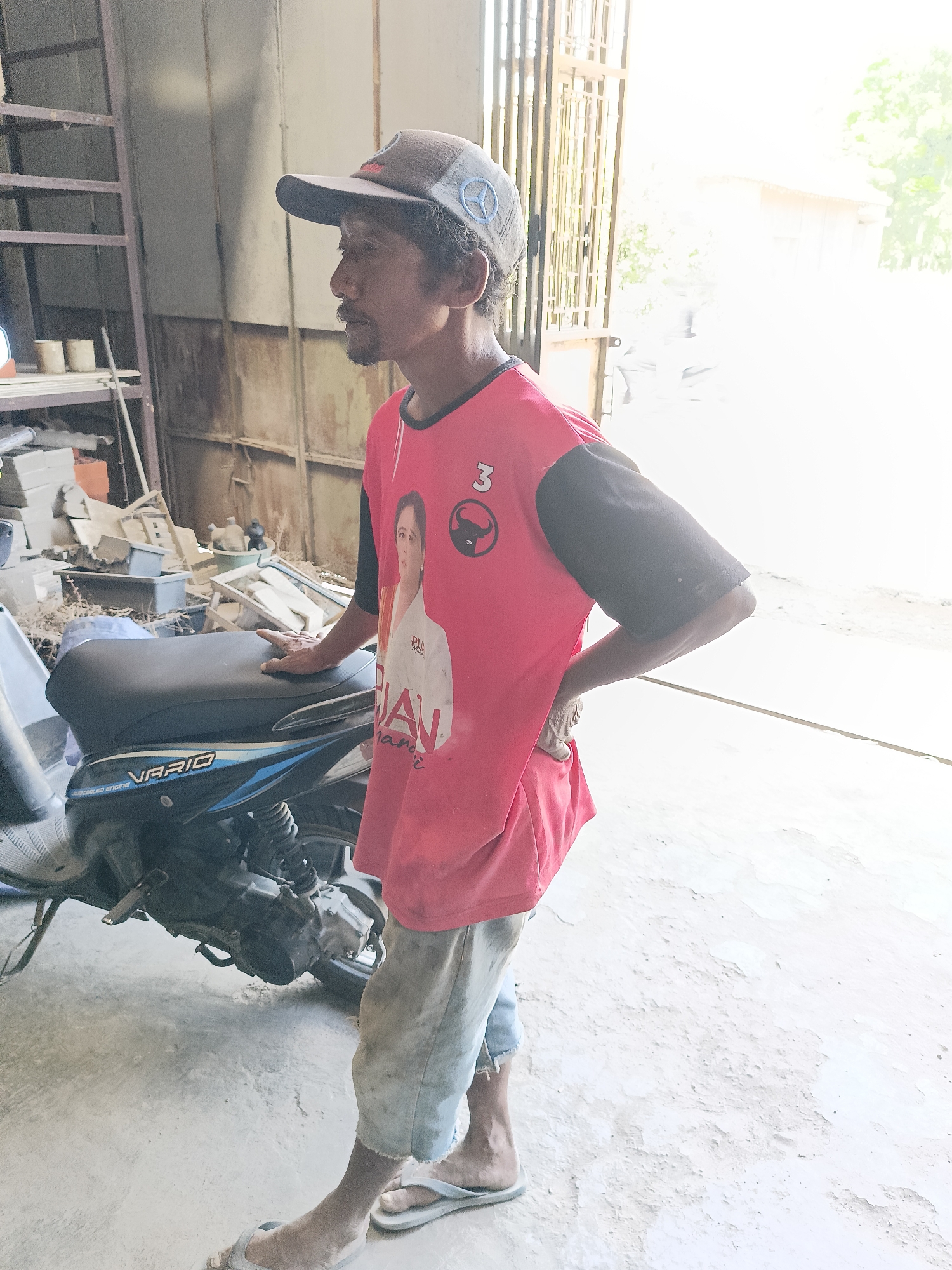A man stands next to a motorcycle in what appears to be a workshop or storage area in Sukorini, Indonesia. The man is wearing a red t-shirt with a picture of a woman and a logo, a baseball cap, and light-colored, torn pants. He has his right hand on the seat of the motorcycle. The motorcycle is dark-colored with blue accents and the brand name "VARIO" is visible. The environment is cluttered with various items, including what looks like building materials, tools, and shelves. The lighting suggests it is daytime, with bright light coming from an opening, possibly a doorway or window, in the background.