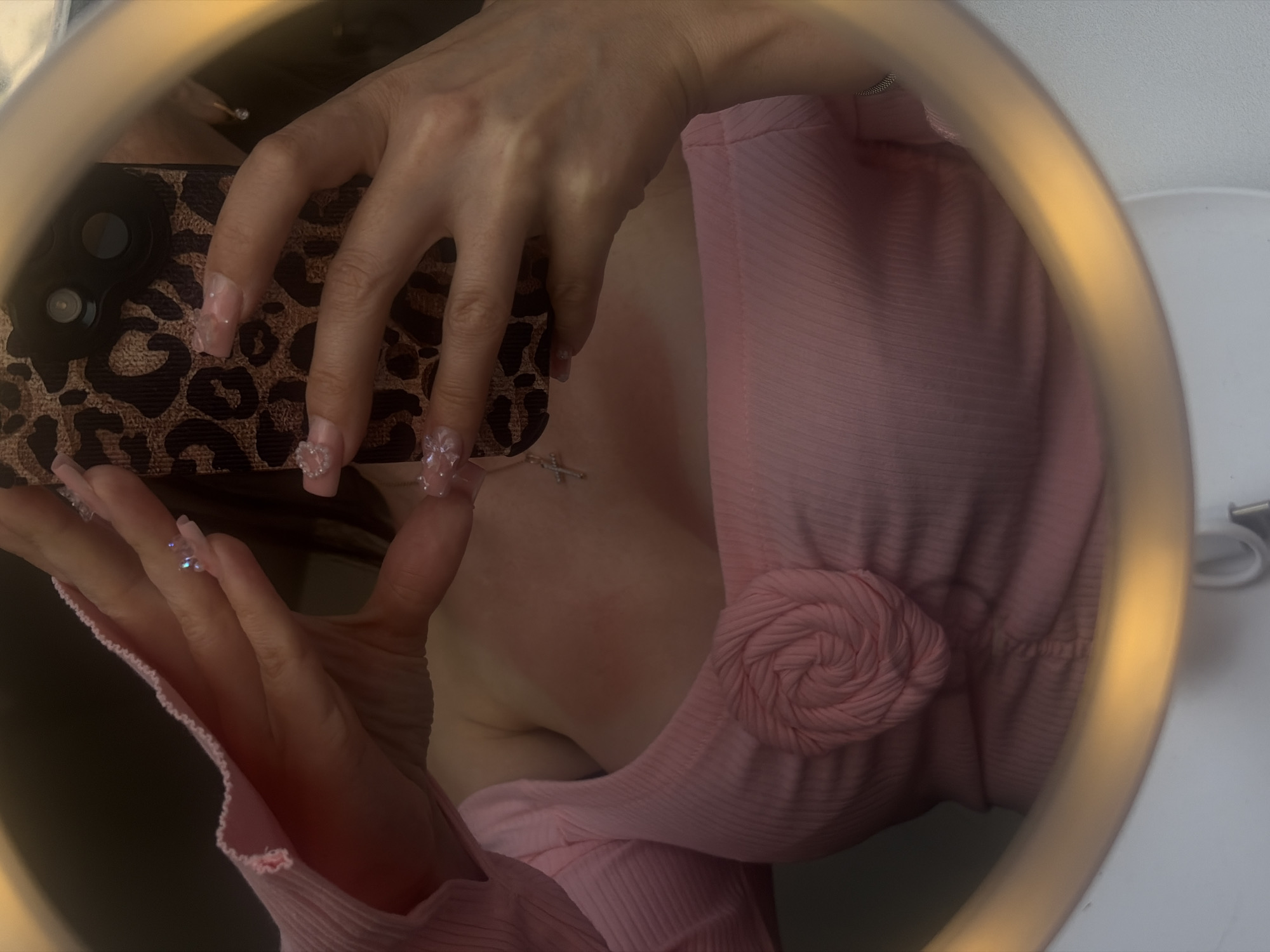 A person is taking a selfie in a mirror. The person is wearing a pink ribbed long-sleeved top with a rose detail in the center. Their hands are visible, holding a phone with a leopard print case. The phone's camera is pointed towards the mirror, capturing the reflection. The person has long, light-colored nails with decorations. There appears to be some redness or blush on the chest area of the person. The background is blurred, but a ring light is visible around the edge of the mirror, suggesting the photo was taken in a well-lit environment. No specific location in the city of Unknown, Russia can be identified.